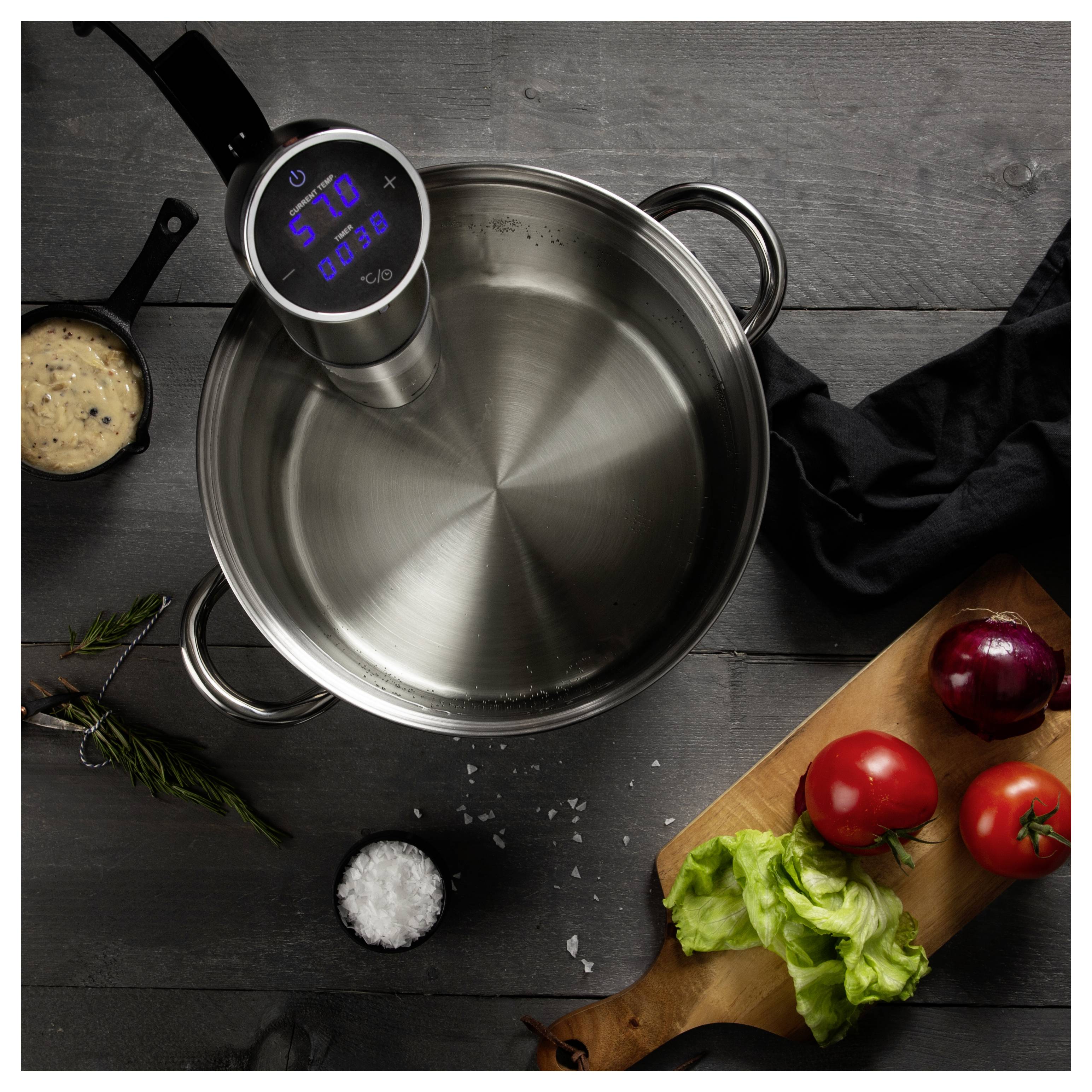 Sous-vide-Garer mit Klammer an einem Edelstahltopf auf einer dunklen Arbeitsfläche befestigt, mit Gemüse und Zutaten, die drumherum angeordnet sind.