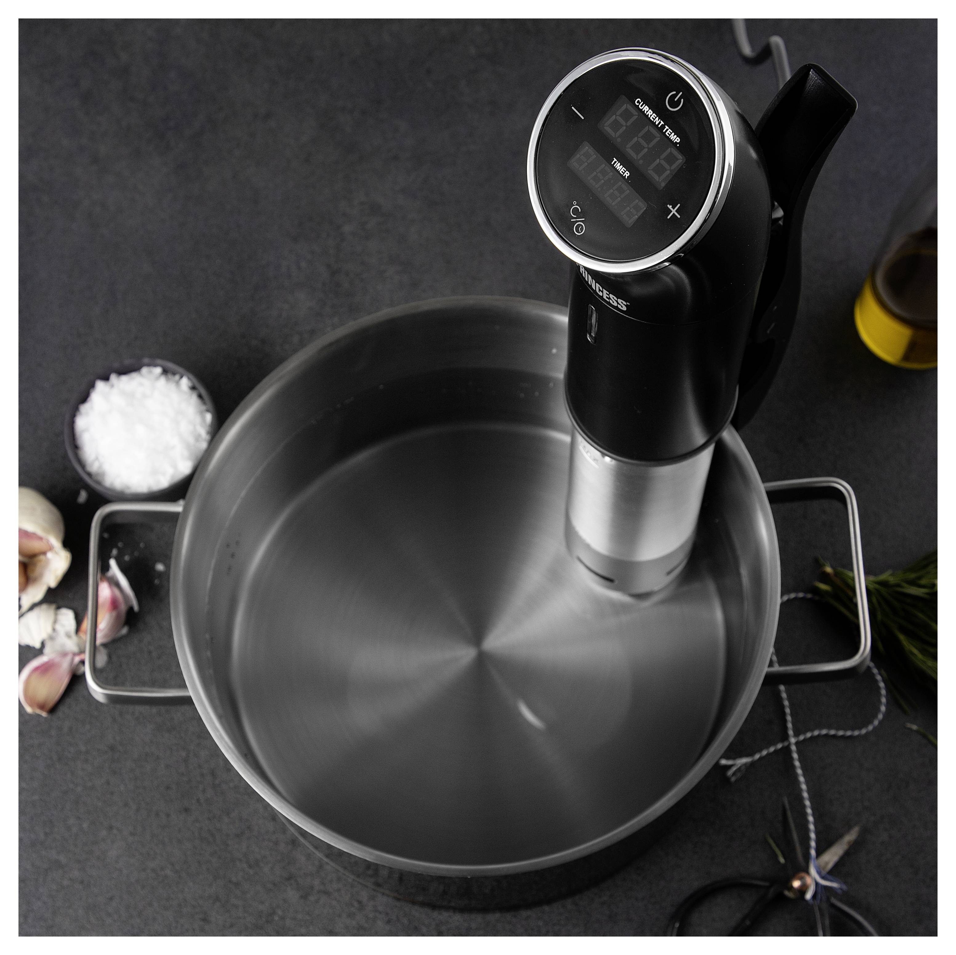 Immersionszirkulator erhitzt Wasser in einem großen Edelstahltopf zum Sous-Vide-Kochen, mit Salz, Knoblauch und Kräutern in der Nähe.
