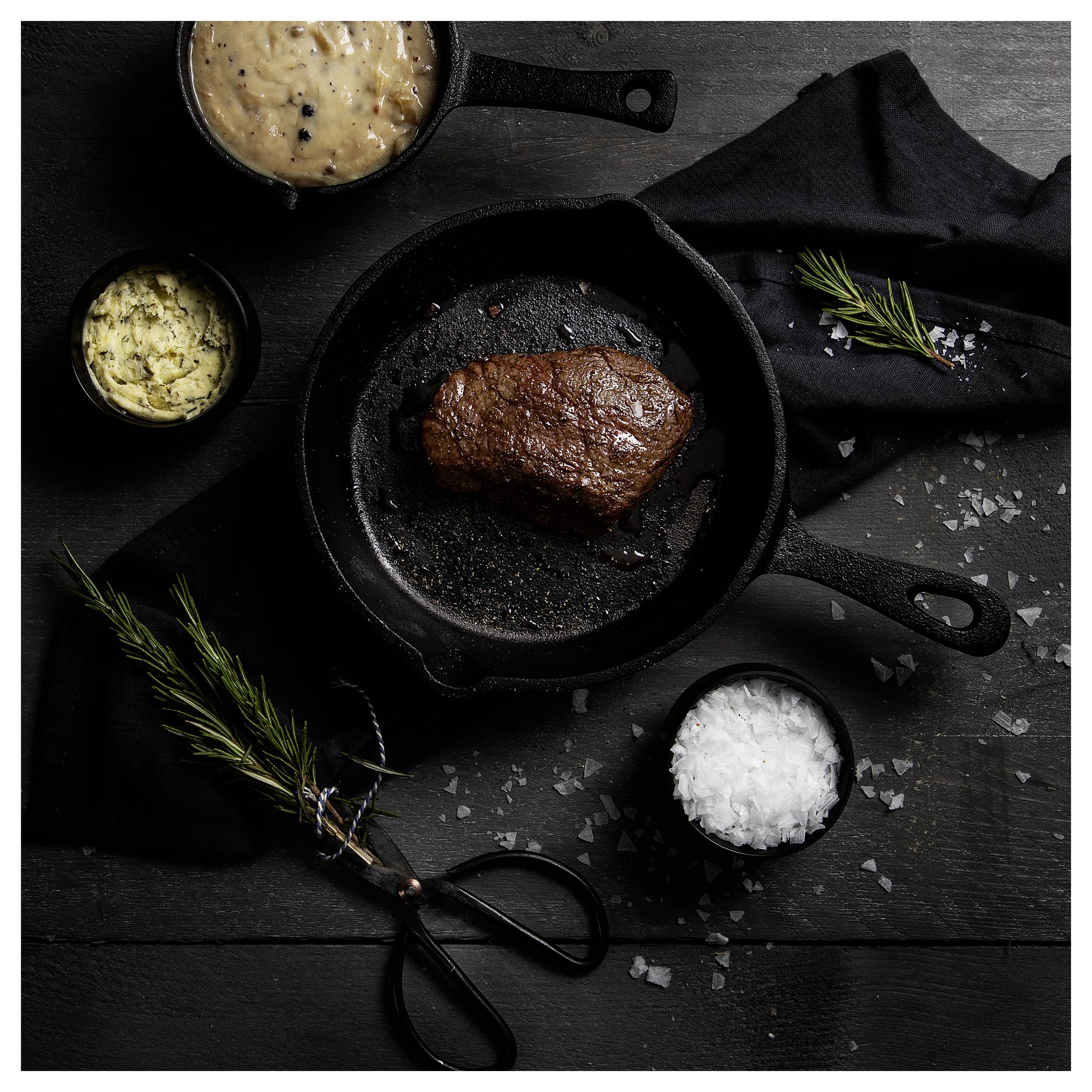 Angebratenes Steak in einer Gusseisenpfanne auf einem dunklen Tisch, umgeben von kleinen Schüsseln mit Sauce, Salz und Rosmarinzweigen.