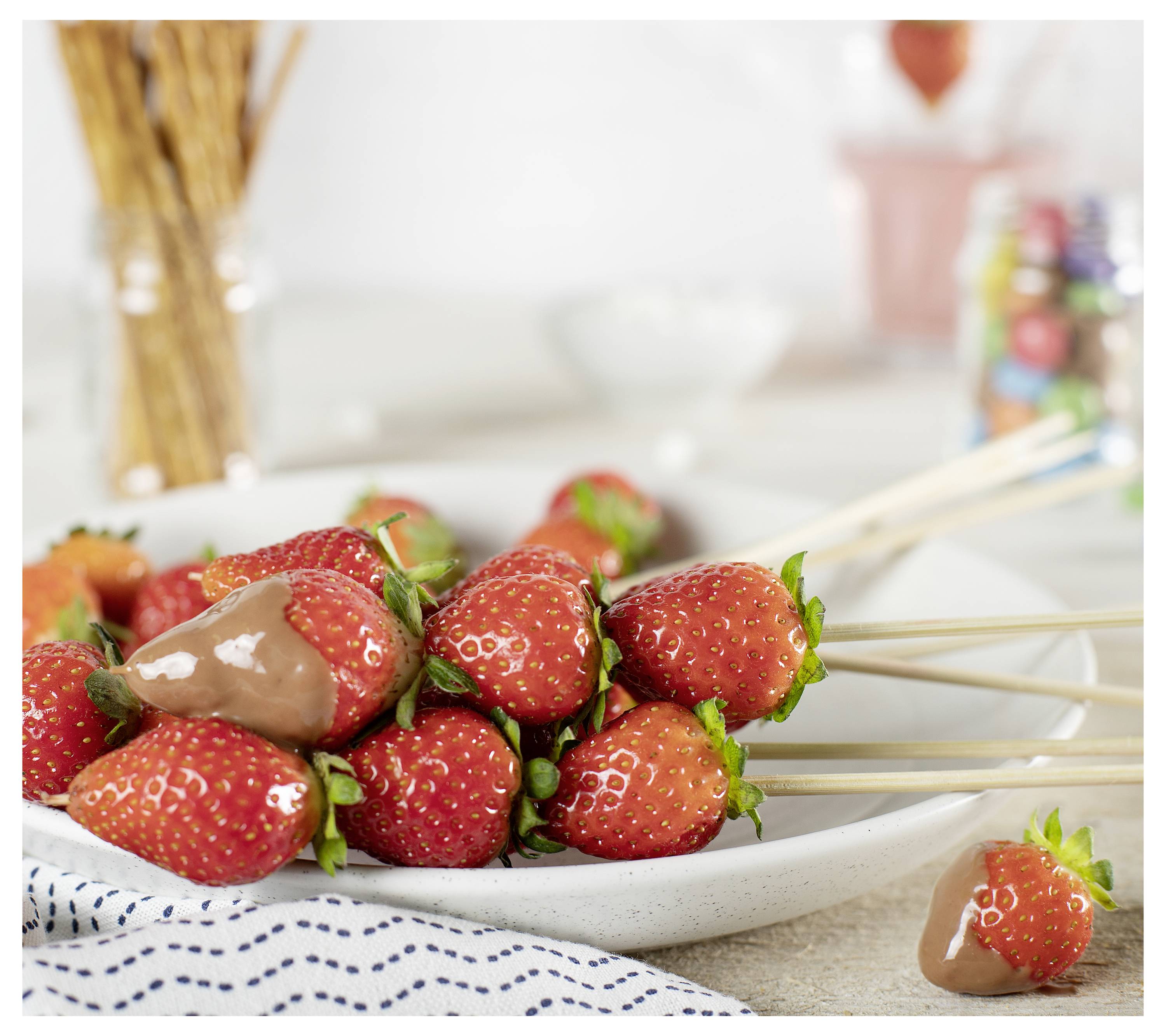Schokoladenüberzogene Erdbeeren auf Holzspießen, aufgehäuft auf einem Teller, mit einer Erdbeere neben dem Teller; Dessert- oder Snack-Präsentation.