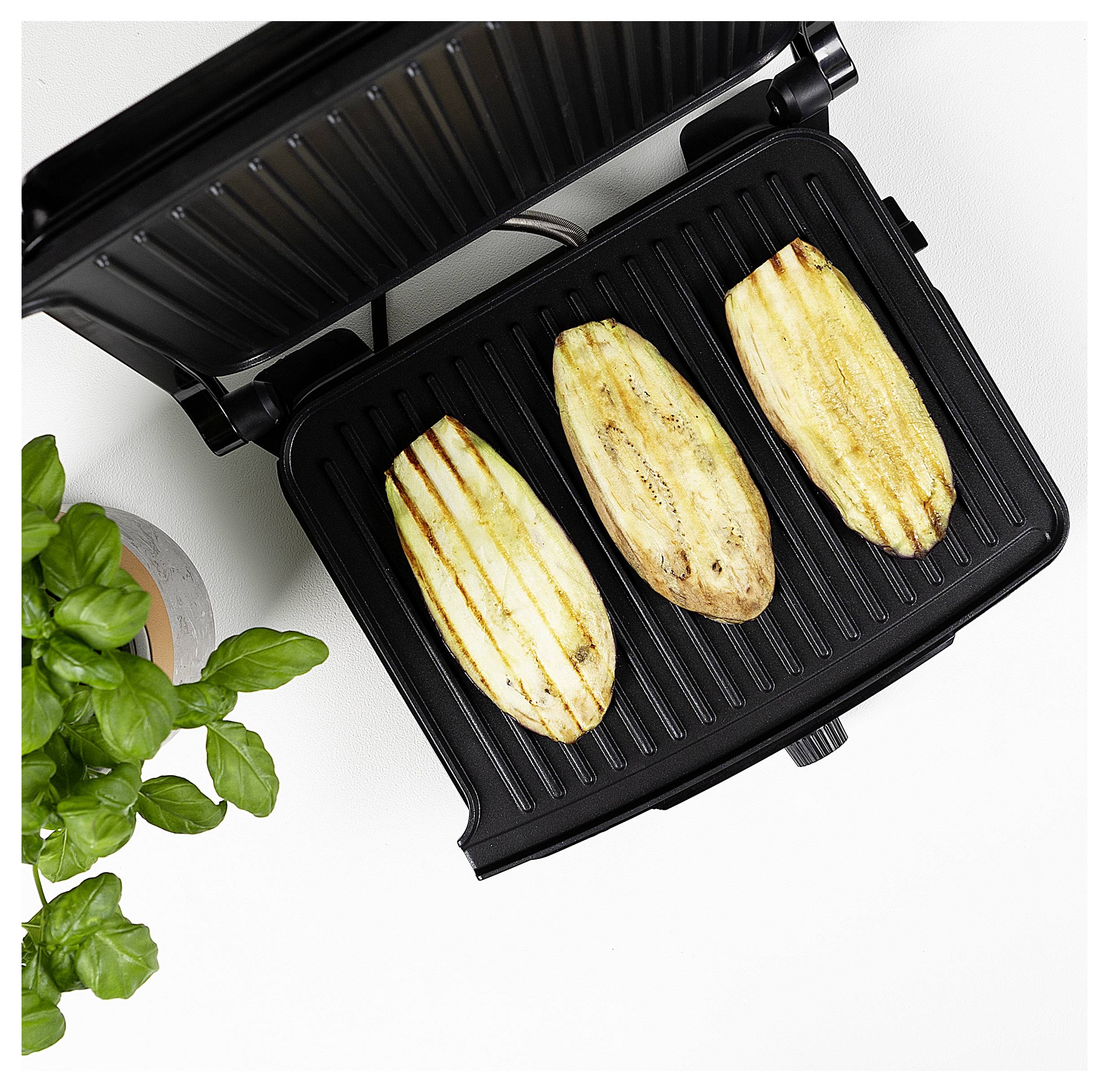 Offener Kontaktgrill mit drei gegrillten Zucchinicheiben und einer Topfpflanze mit Basilikum auf einer weißen Oberfläche.