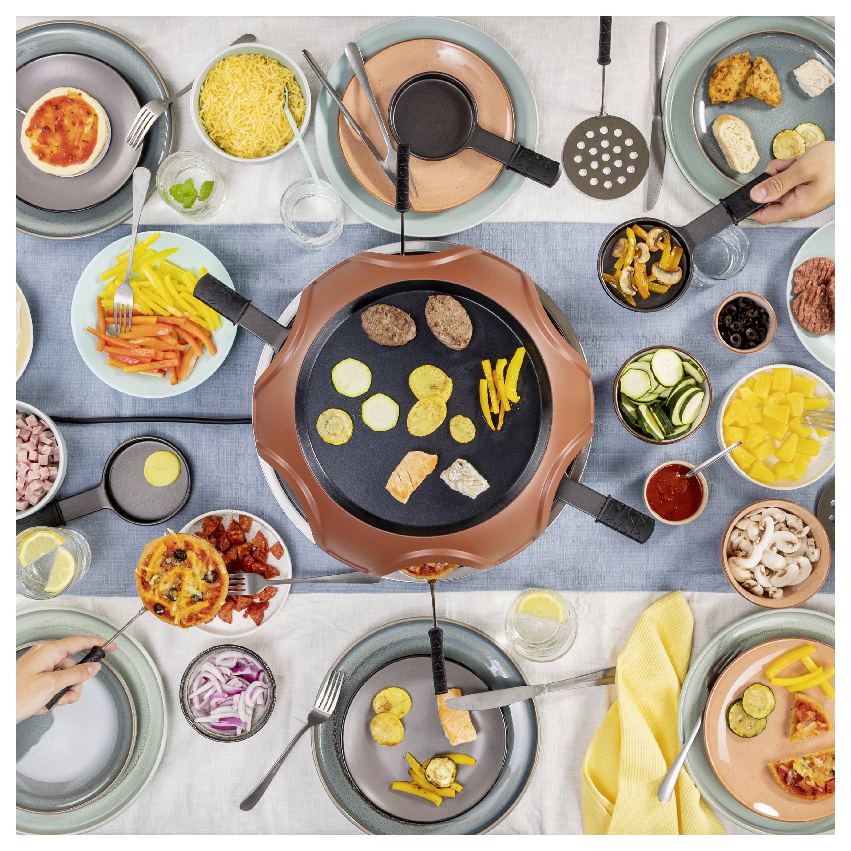 Aufsicht auf ein gemeinschaftliches Fondue-Essen mit Pfannen mit geschmolzenem Käse und Schokolade, umgeben von verschiedenen Gemüsesorten, Fleisch, Brot und Obst zum Eintauchen.