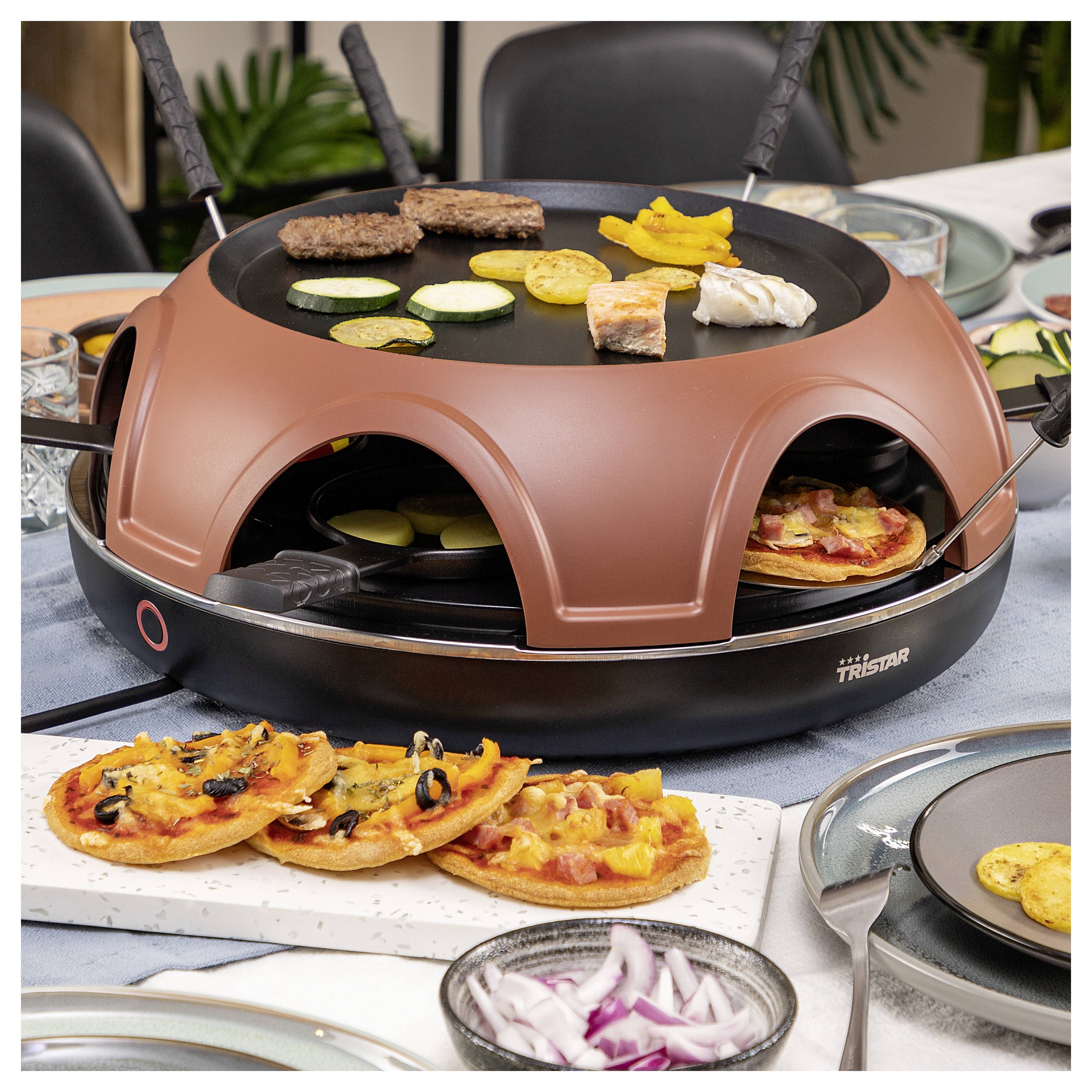 Ein Tischgrill und Raclette-Gerät mit Fleisch, Gemüse, Garnelen und Mini-Pizzas beim Kochen, mit angerichtetem Essen und Zwiebelscheiben rundherum.