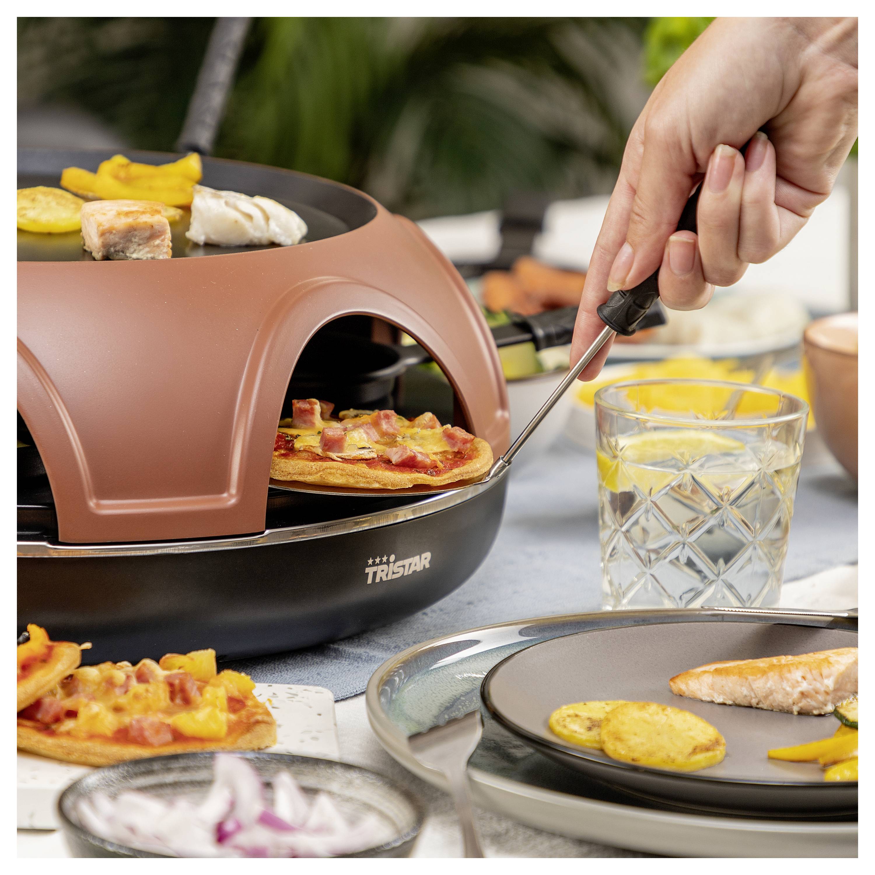 Eine Hand benutzt eine Pizzaschaufel, um eine Mini-Pizza aus einem Tischplatten-Pizzaofen im Innenbereich neben Tellern mit gekochtem Essen und einem Getränk zu entfernen.