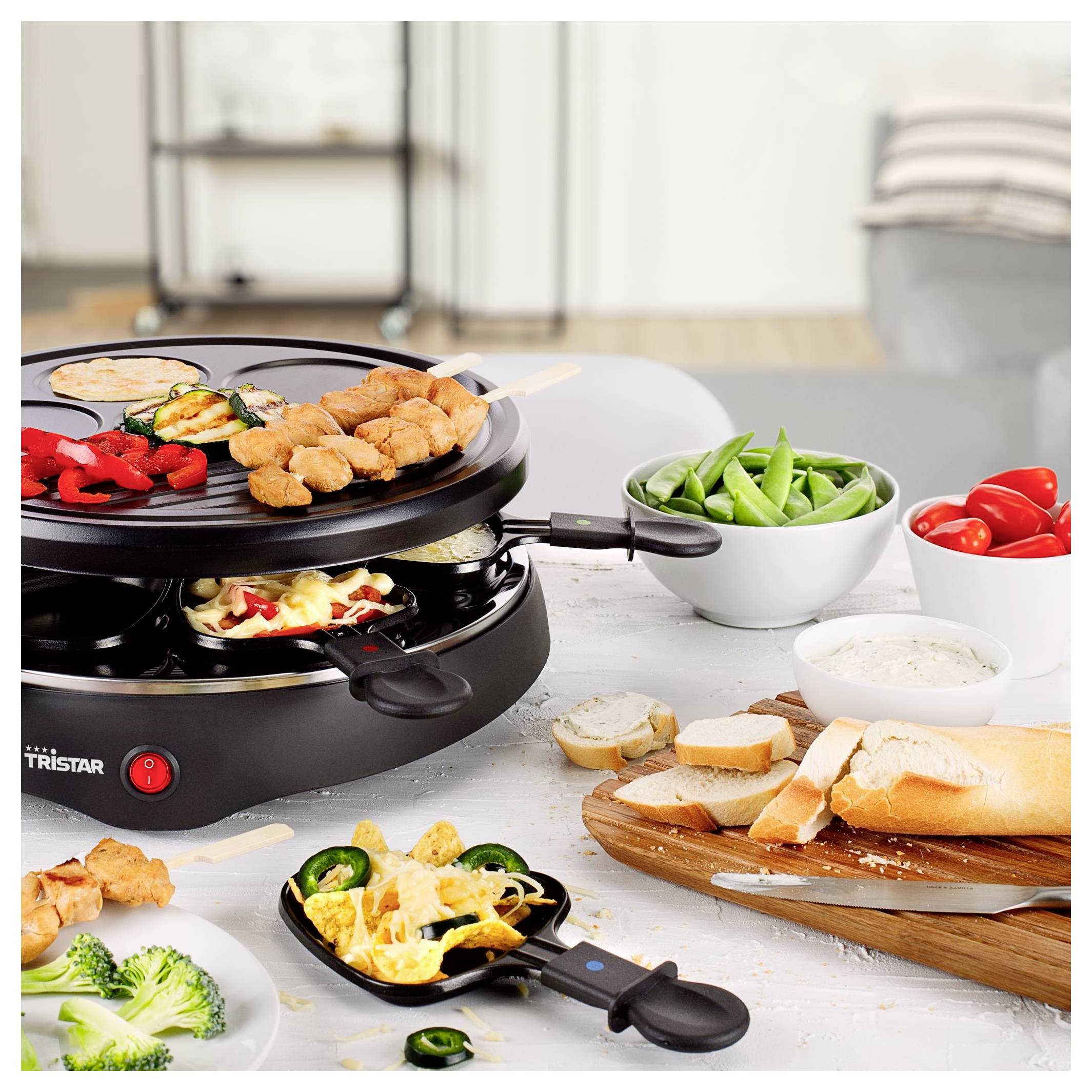 Tragbarer Tischgrill für den Innenbereich mit Spießen aus Fleisch und Gemüse, umgeben von Schüsseln mit Zuckerschoten und Tomaten, Brot, Dip und servierten Beilagen.