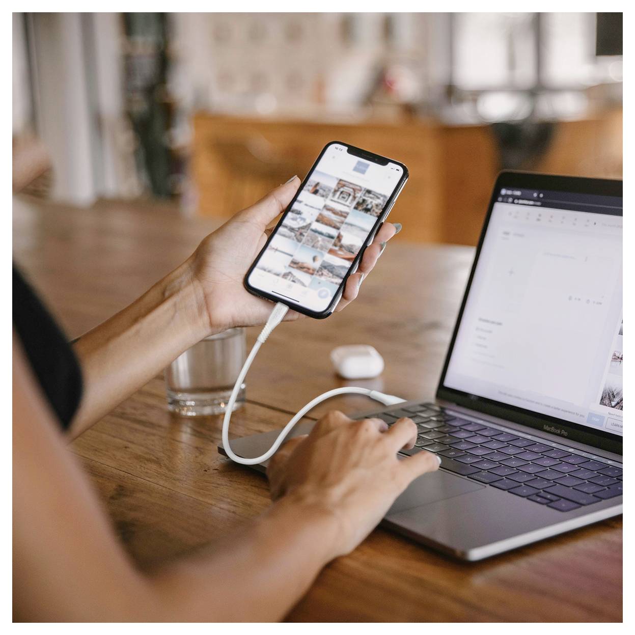 Person, die ein Smartphone per Kabel mit einem Laptop verbunden hat, während sie an einem Holztisch arbeitet, wobei das Telefon eine Fotogalerie anzeigt und der Laptop offen ist.