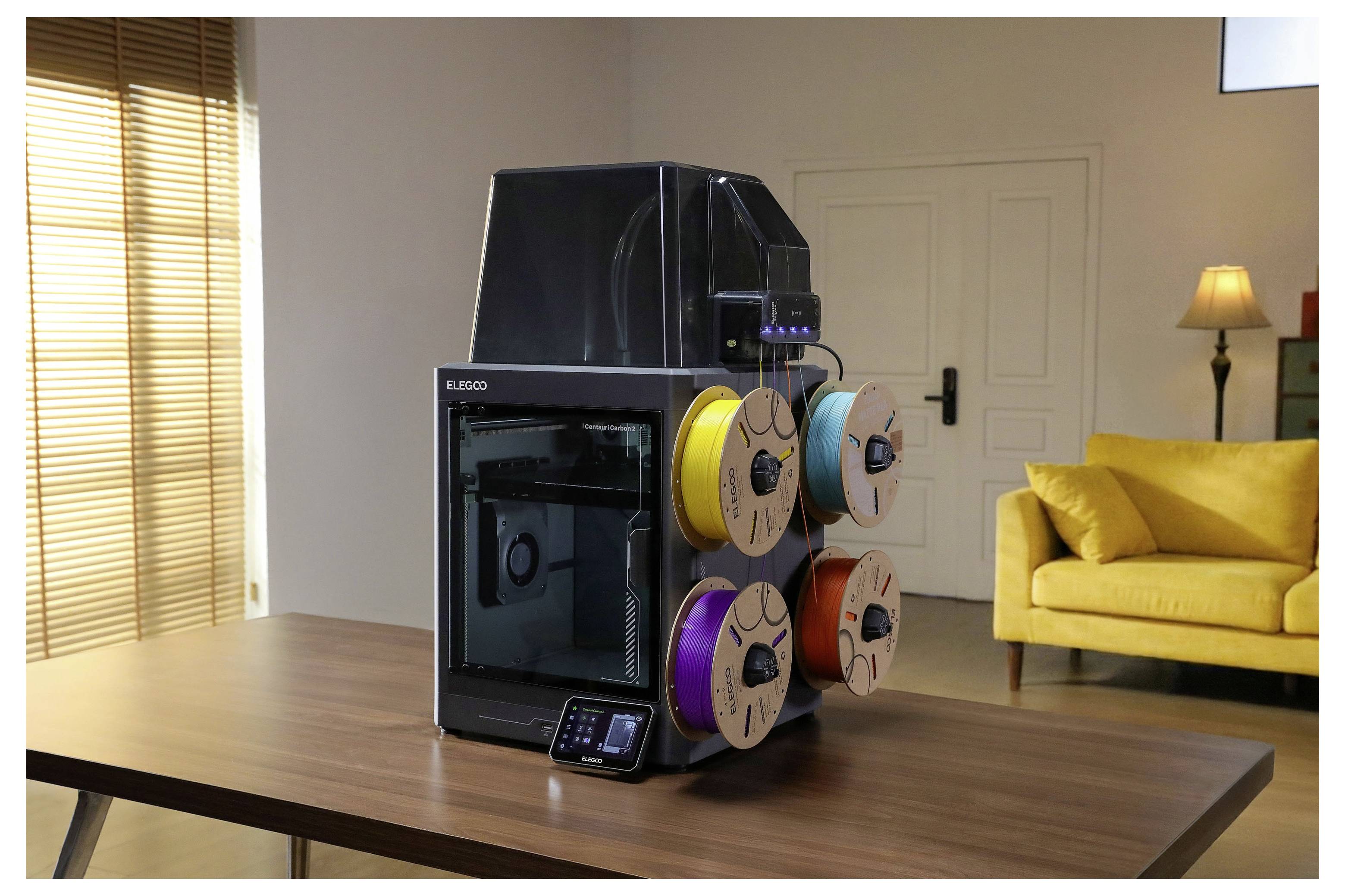 3D-Drucker mit vier Filamentspulen auf einem Holztisch in einem Wohnzimmer, mit einem gelben Sofa und einer Stehleuchte im Hintergrund.