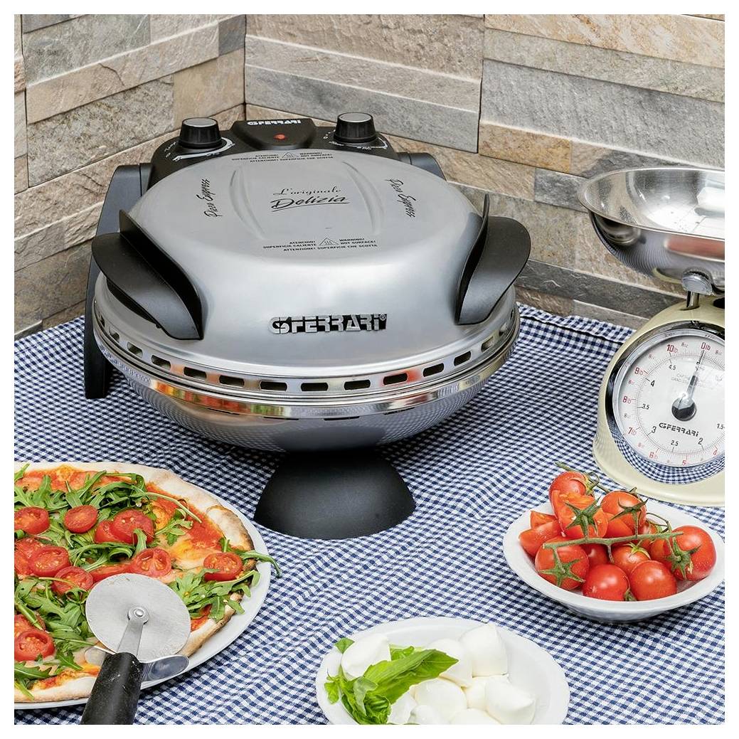 Ein Tischgrill an einer Buffetstation brät Speisen neben einer Waage, einer Pizza, Kirschtomaten und Mozzarella auf einem karierten Tuch.