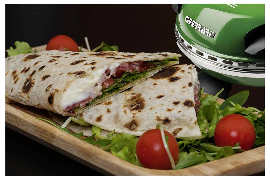 Gegrilltes Wrap-Sandwich, in der Mitte durchgeschnitten auf einem Holzbrett, gefüllt mit geschmolzenem Käse und Grünzeug, mit Kirschtomaten neben einer grünen Panini-Presse.
