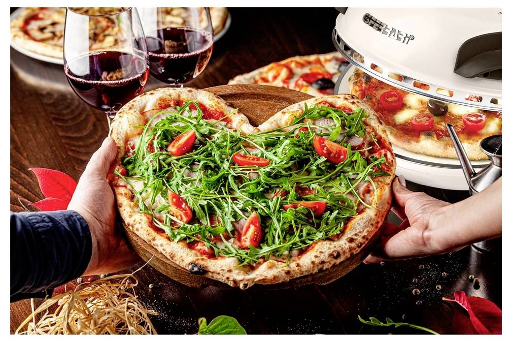 Zwei Hände präsentieren eine Pizza mit Rucola und Kirschtomaten neben Gläsern Rotwein und einem Arbeitsplatten-Pizzaofen.