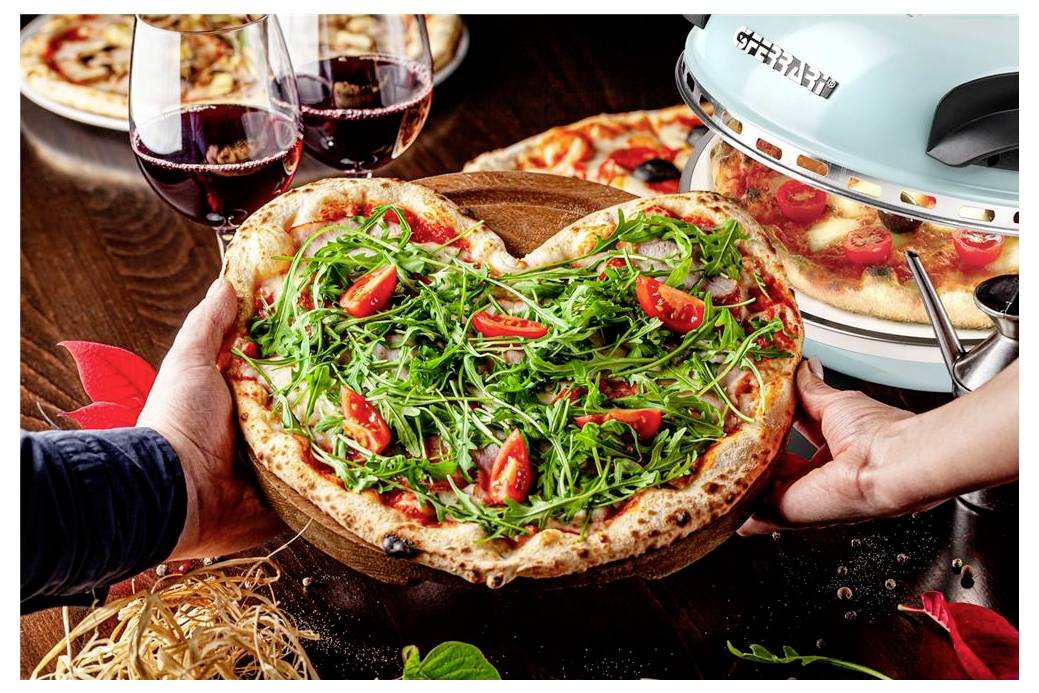 Hände halten eine herzförmige Rucola- und Tomatenpizza auf einem Holzbrett, mit Weingläsern und einem Pizzaofen im Hintergrund.