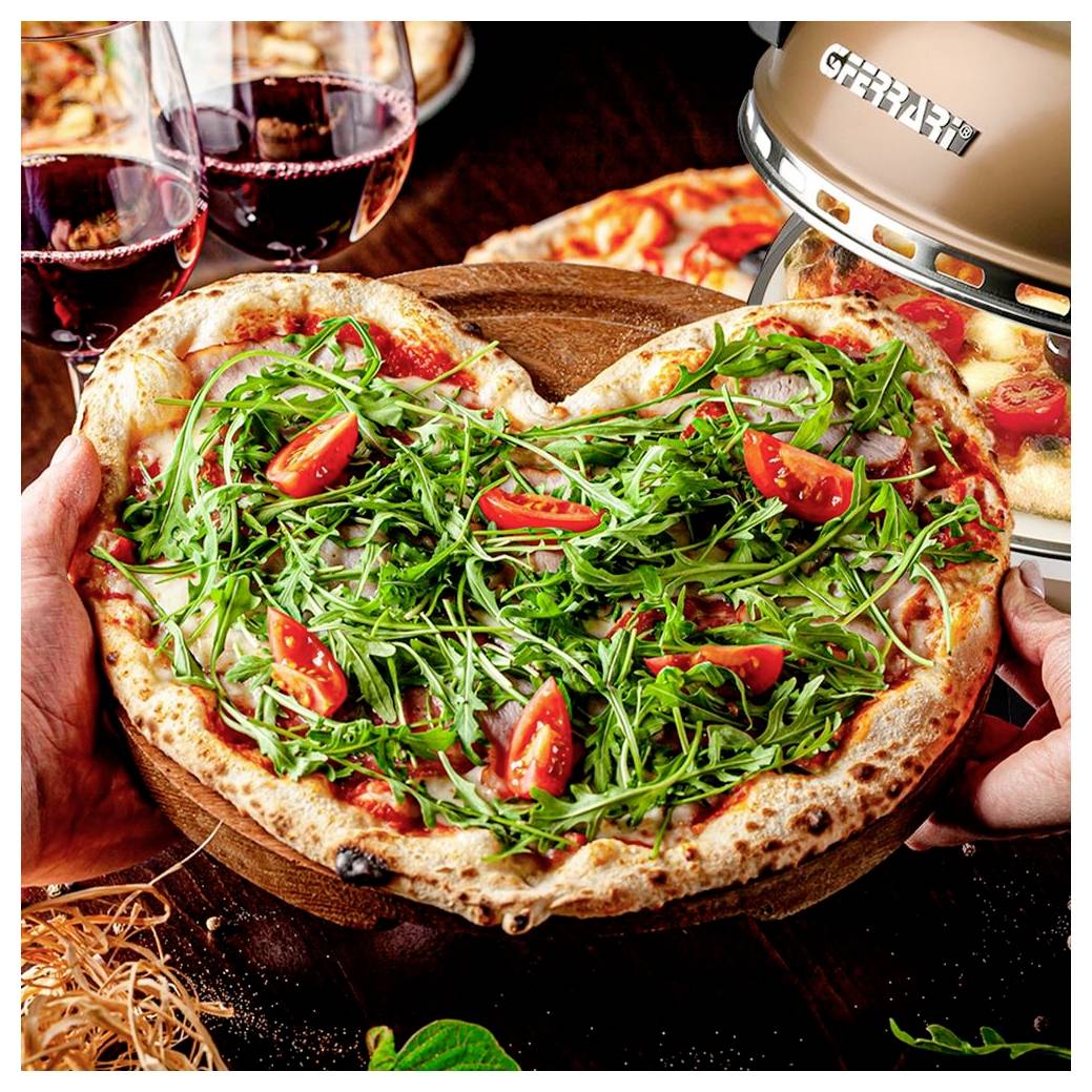 Herzförmige Pizza mit Rucola und Kirschtomaten, die hochgehalten wird, im Hintergrund Weingläser und ein Tischgrill.