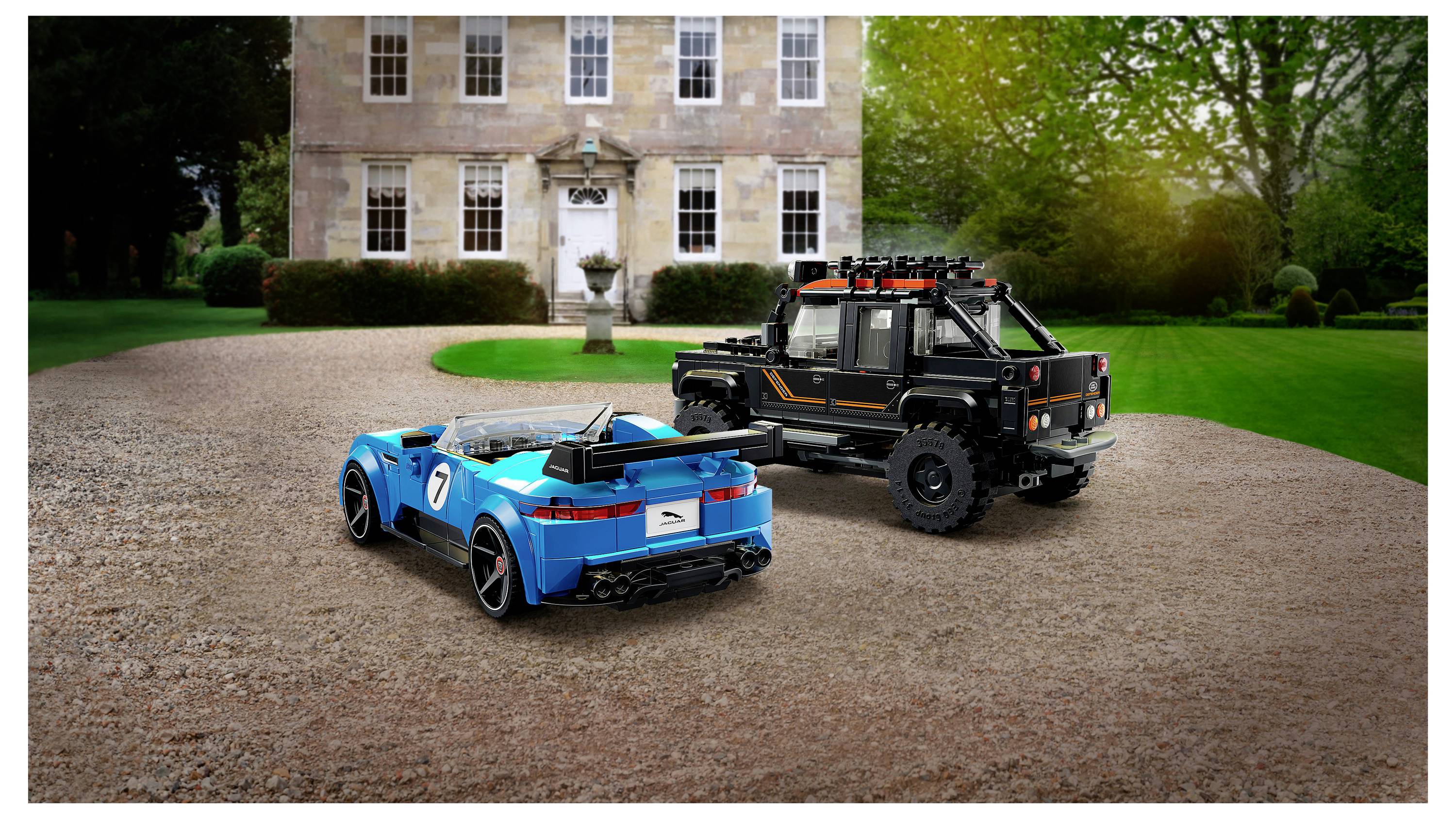 Zwei LEGO-ähnliche Geländefahrzeuge, ein blauer Sportwagen und ein schwarzer Nutzwagen, parken auf einer Kiesauffahrt vor einem großen Haus.