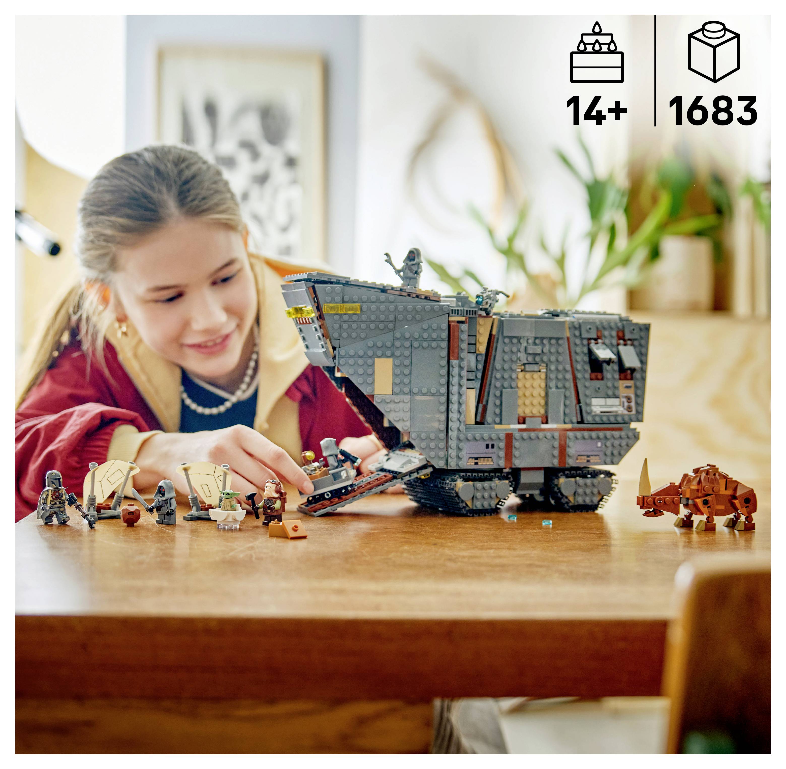 Ein Mädchen spielt mit einem großen grauen LEGO-Raumschiffmodell auf einem Tisch, mit mehreren kleinen Minifiguren und einem orangefarbenen Fahrzeug in der Nähe.