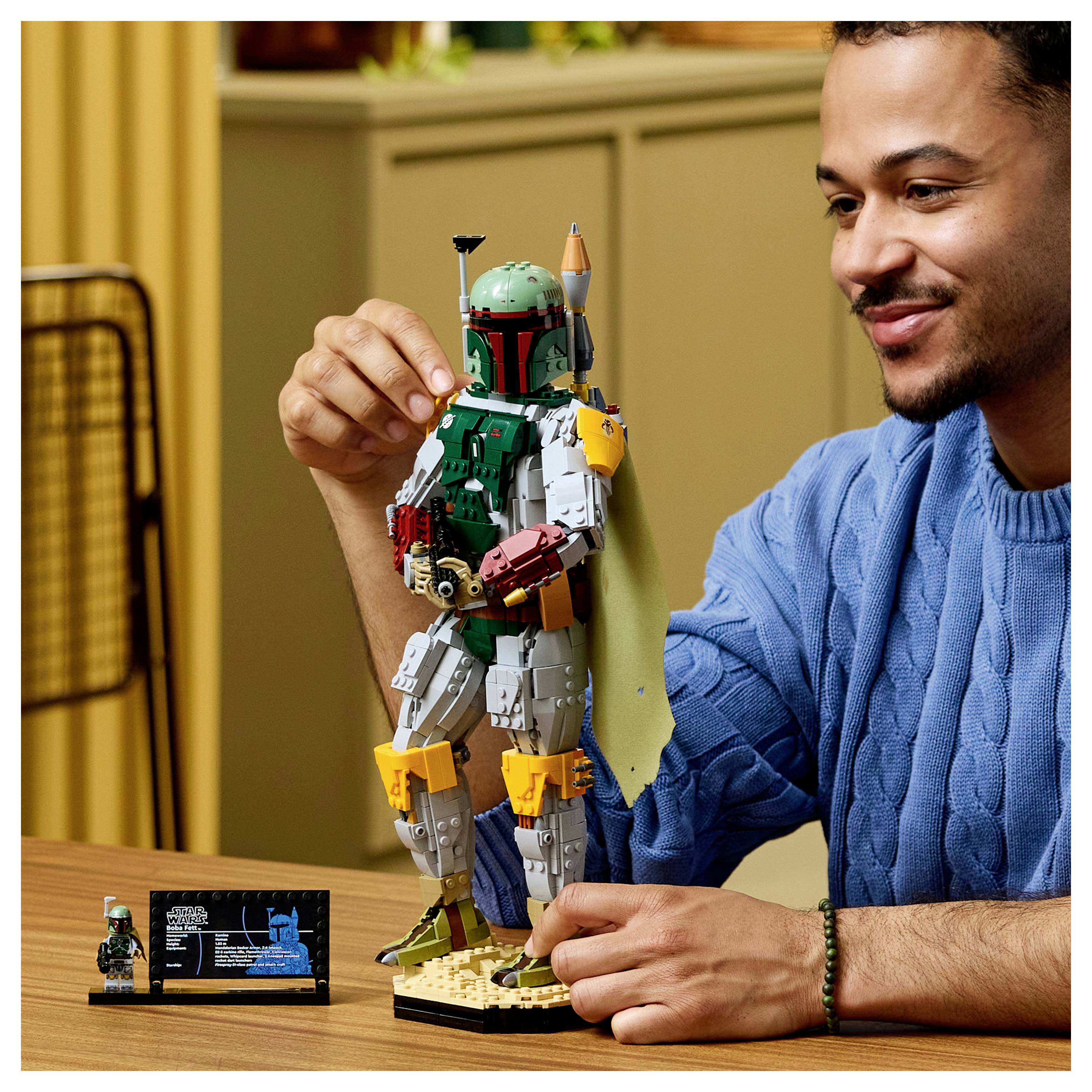 Ein Mann in einem blauen Hemd präsentiert eine LEGO-Boba-Fett-Figur auf einem Tisch und hält sie aufrecht neben ihrem Ausstellungsschild.