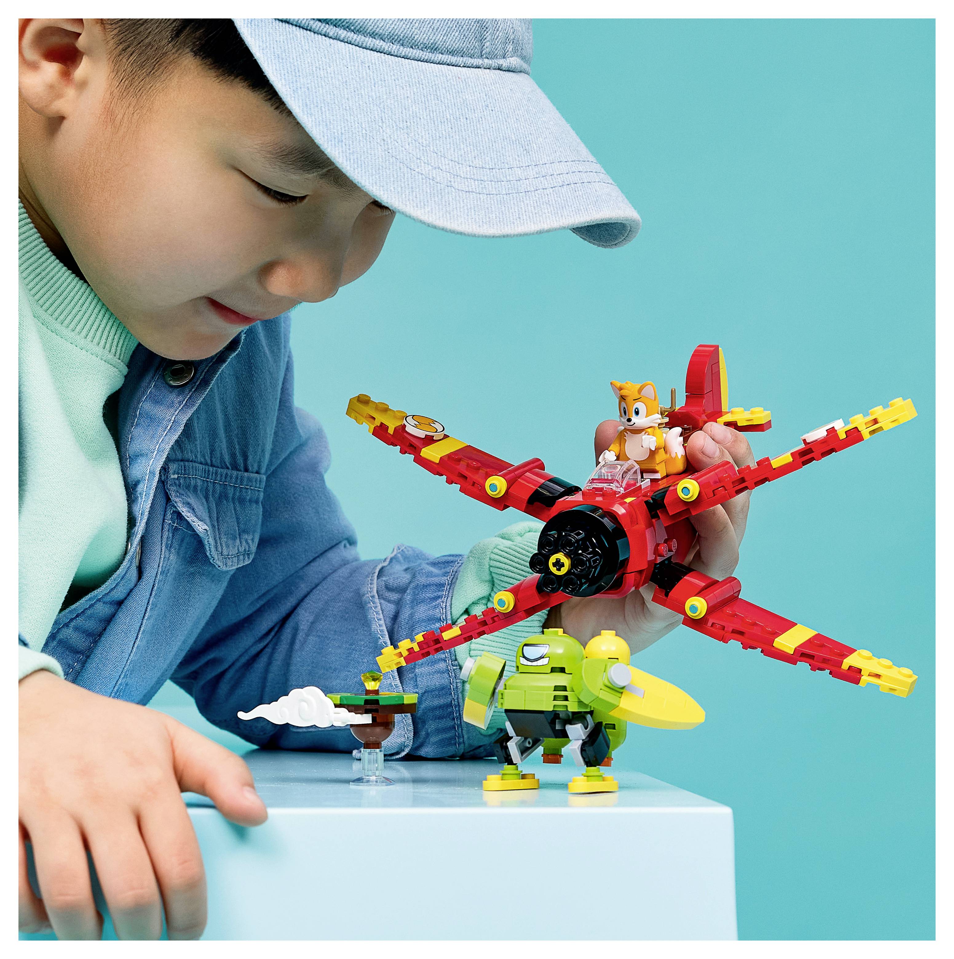 Kleines Kind in einer Mütze, das mit bunten Spielzeugflugzeugen und Figuren auf einem Tisch vor einem blaugrünen Hintergrund spielt.