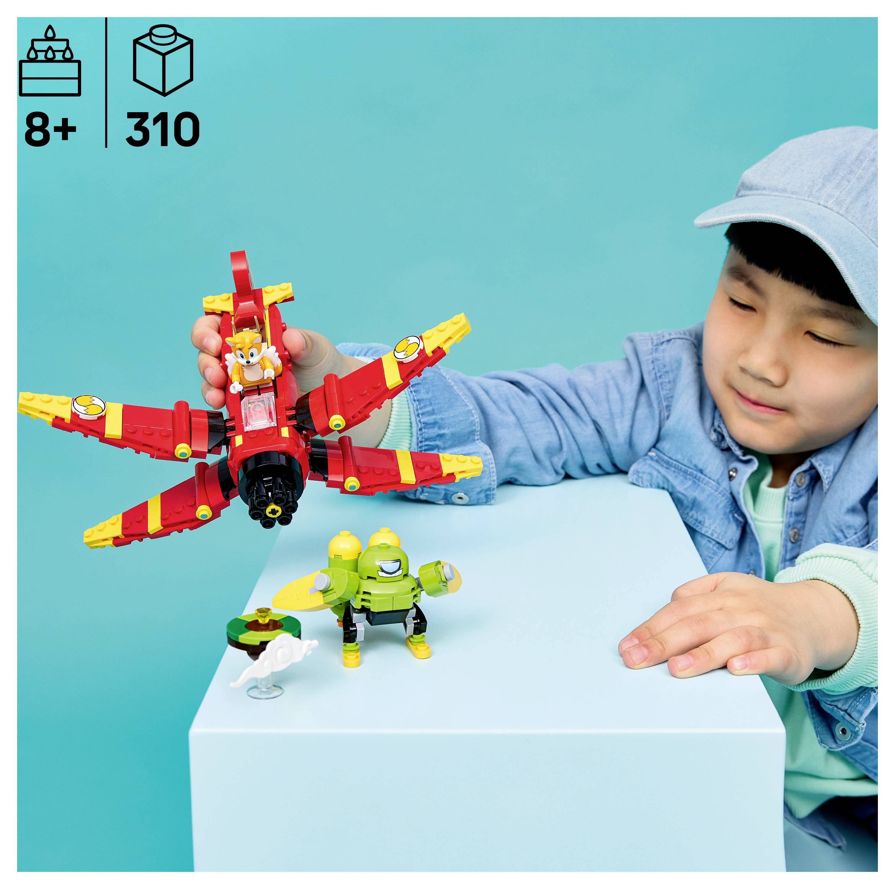 Kleiner Junge spielt mit einem rot-gelben LEGO-Flugwesen und kleinen grünen Figuren auf einem weißen Block, mit „8+