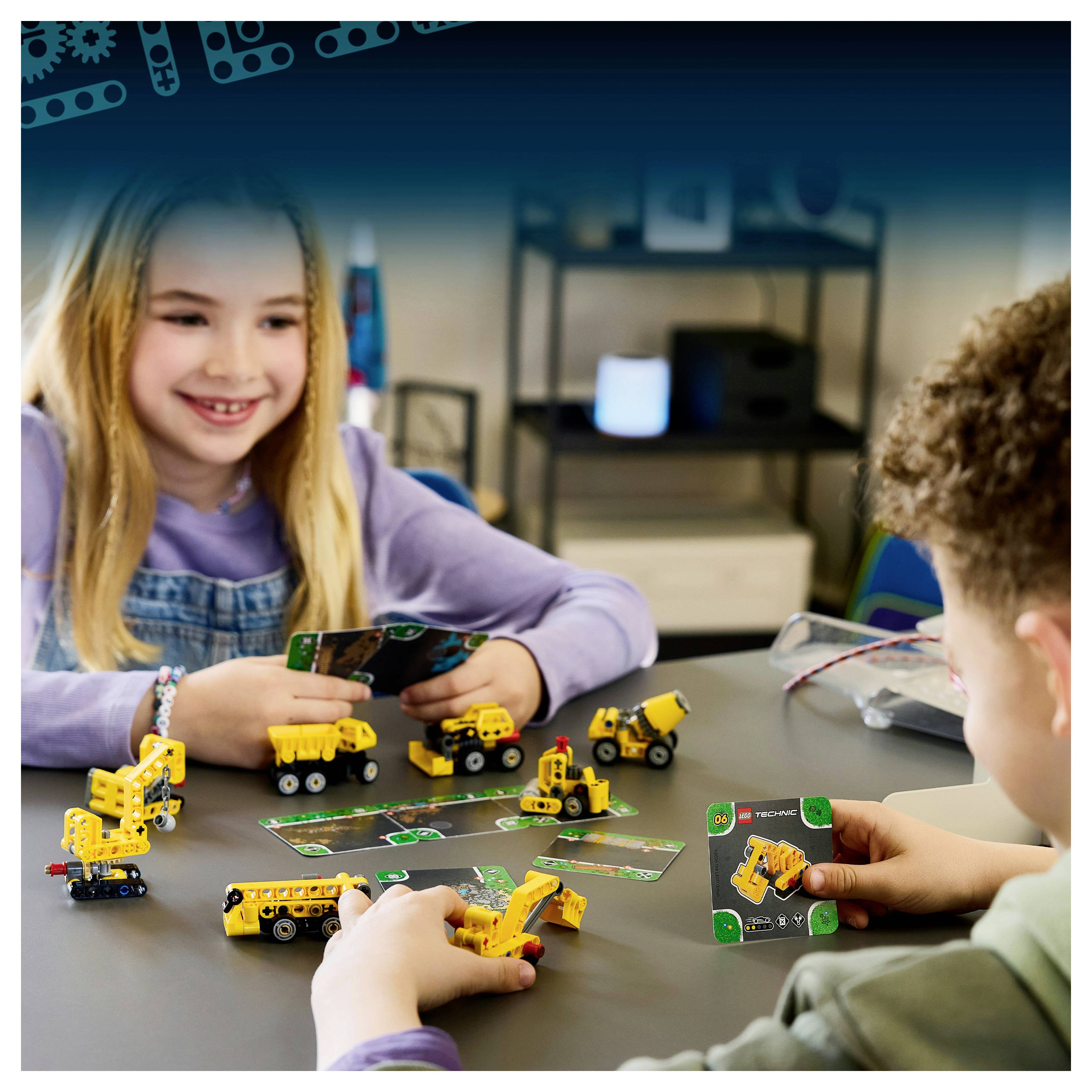 Zwei Kinder spielen an einem Tisch in einem Klassenzimmer oder Spielzimmer mit kleinen gelben Spielzeugbaufahrzeugen und Spielkarten.