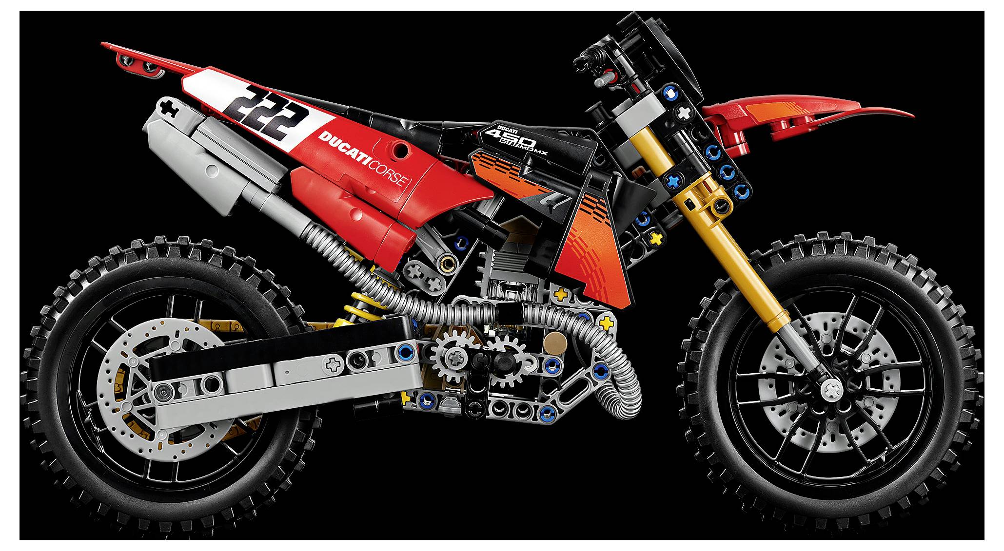Seitenansicht eines roten LEGO-Ducati-DesertX-ähnlichen Geländemotorrad-Modells mit sichtbarem Motor, Aufhängung und Knobby-Reifen auf schwarzem Hintergrund.