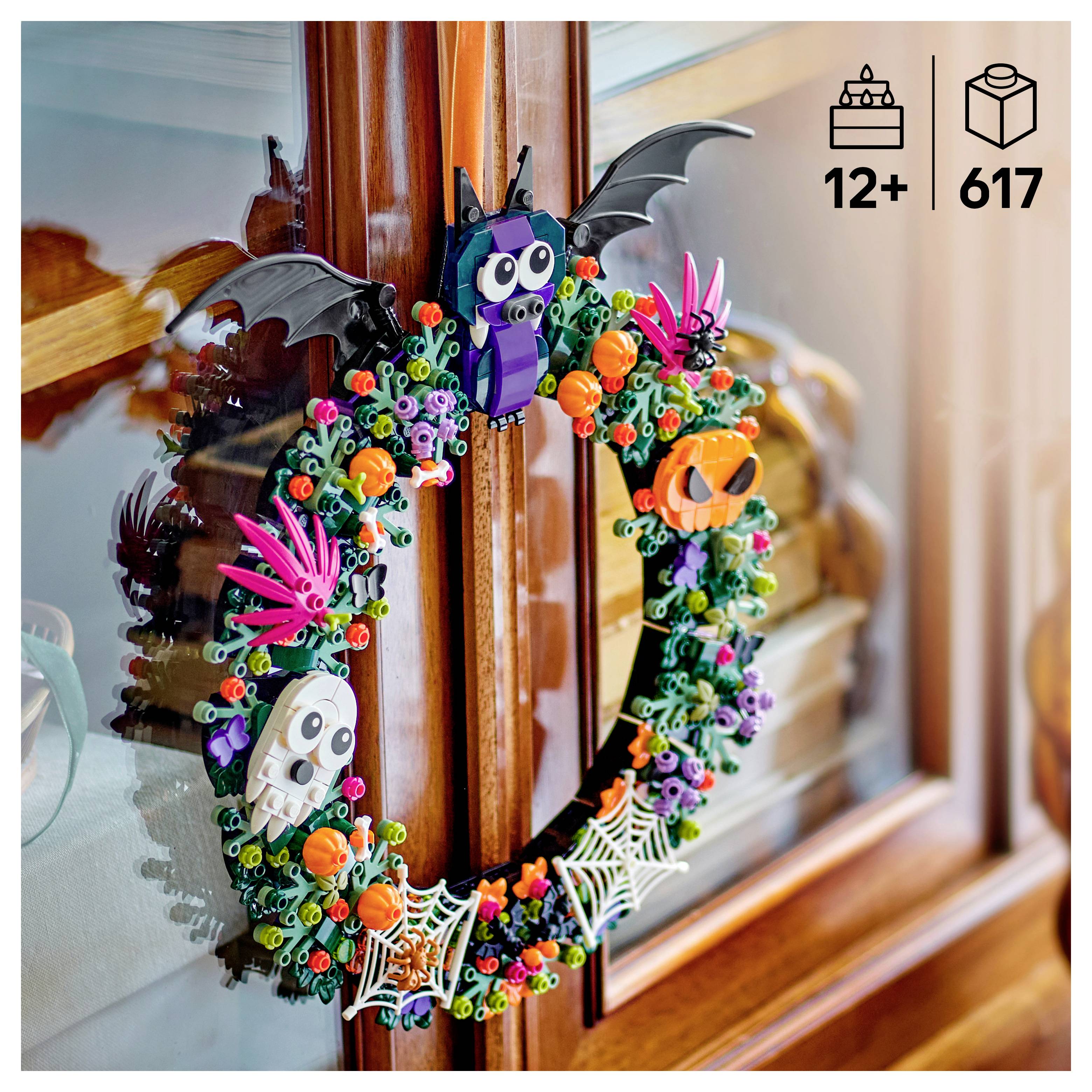 Ein bunter LEGO-Halloweenkranz mit Fledermäusen, Schädeln, Kürbissen, Spinnen und Blumen hängt an einem hölzernen Fensterrahmen.