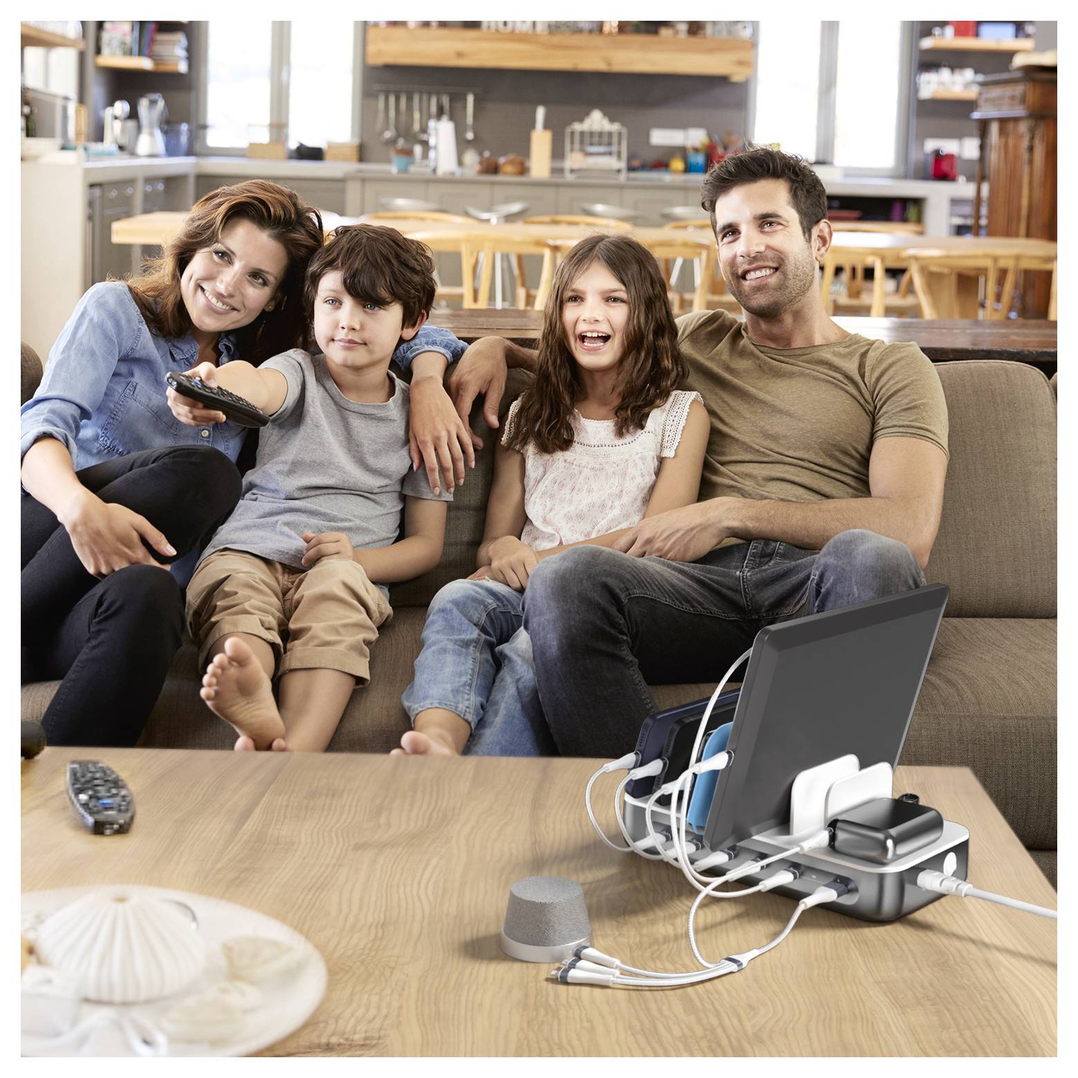 Eine vierköpfige Familie sitzt auf einem Sofa und schaut Fernsehen, wobei sich eine Fernbedienung und aufladende Elektronikgeräte auf dem Couchtisch im Wohnzimmer befinden.