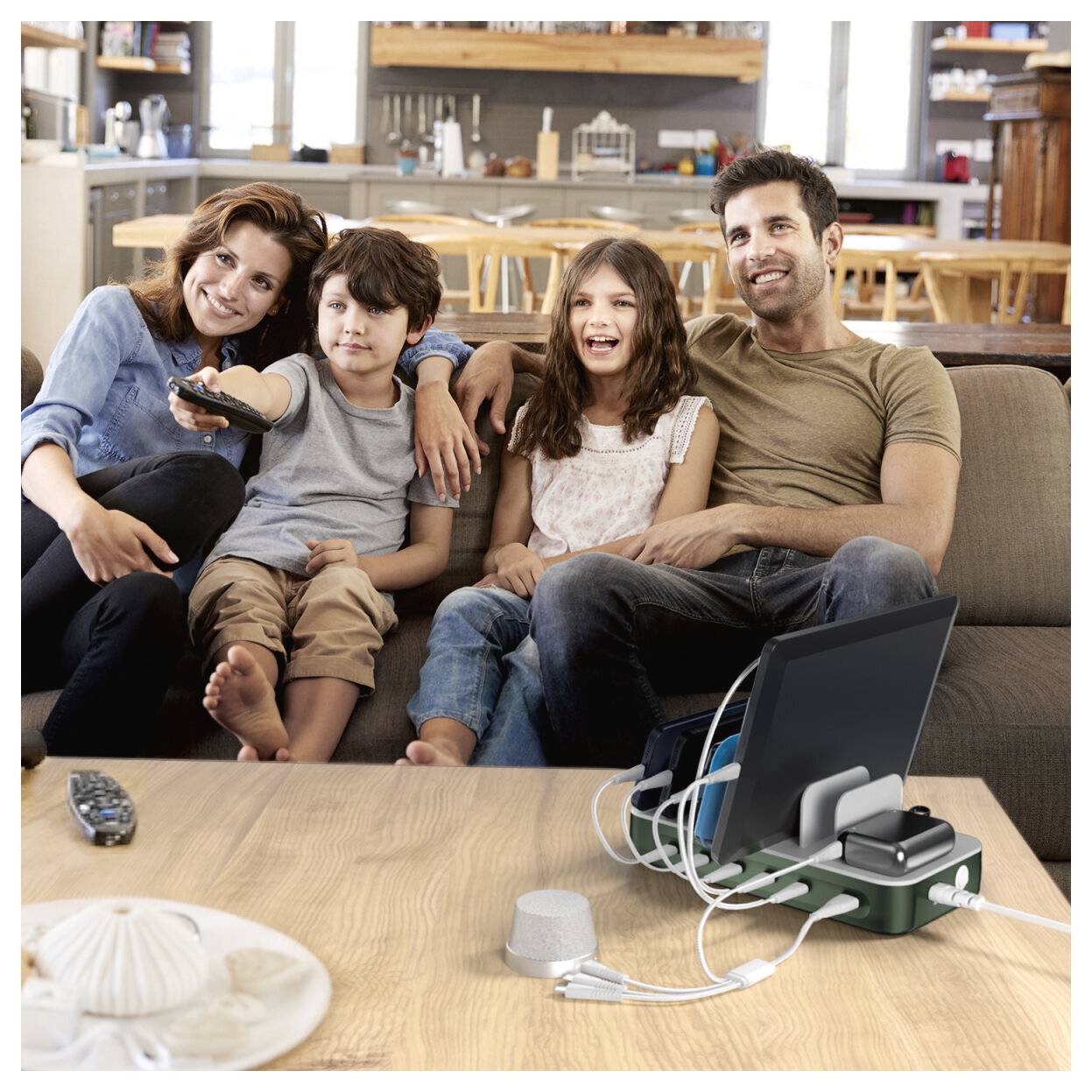 Eine vierköpfige Familie sitzt auf einem Sofa im Wohnzimmer und schaut zusammen fern, während ein Laptop und Ladekabel auf dem Couchtisch liegen.