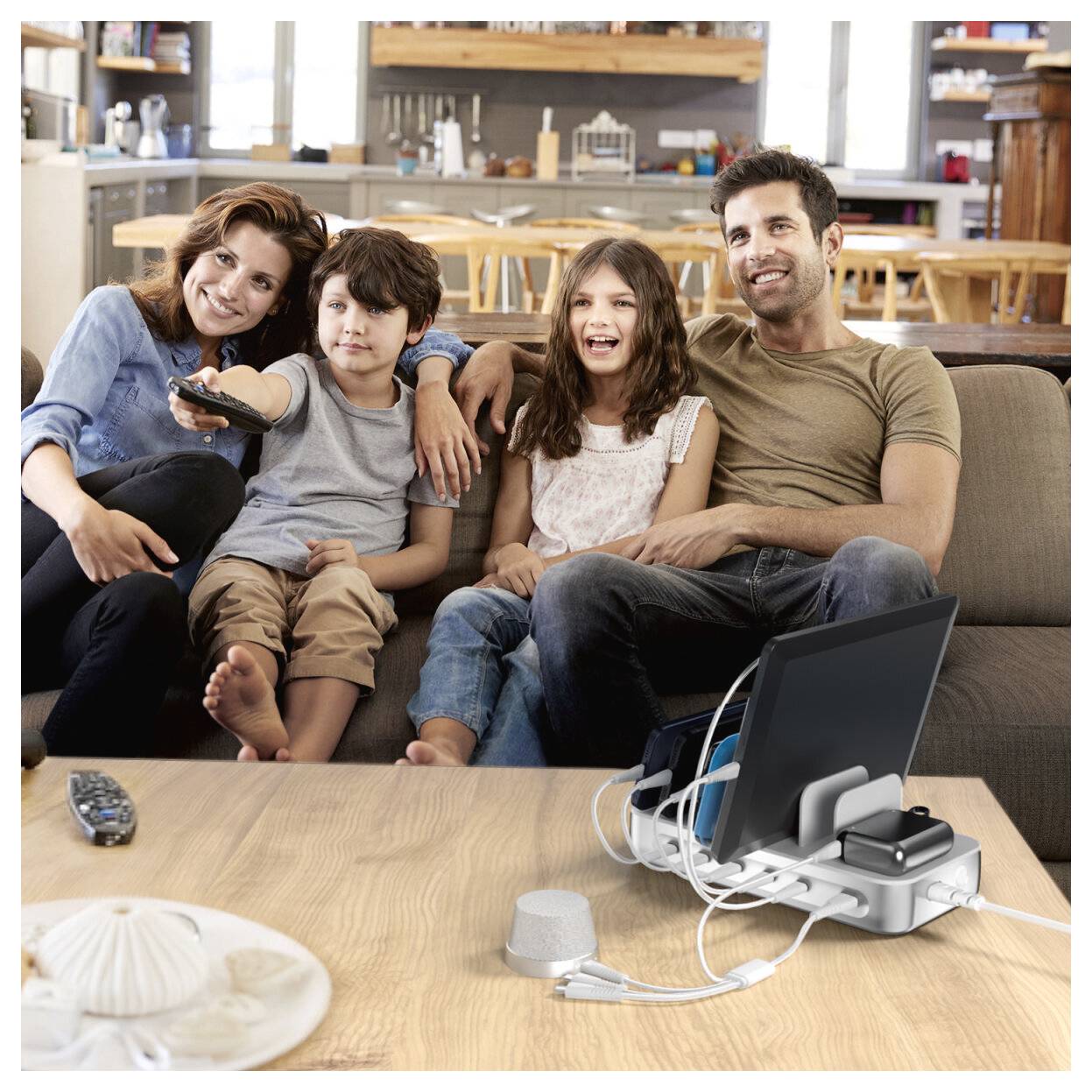 Familie von vier Personen sitzt gemeinsam auf einem Sofa und schaut im Wohnzimmer fern, mit einer Fernbedienung auf dem Tisch und Mediengeräten, die davor angeschlossen sind.