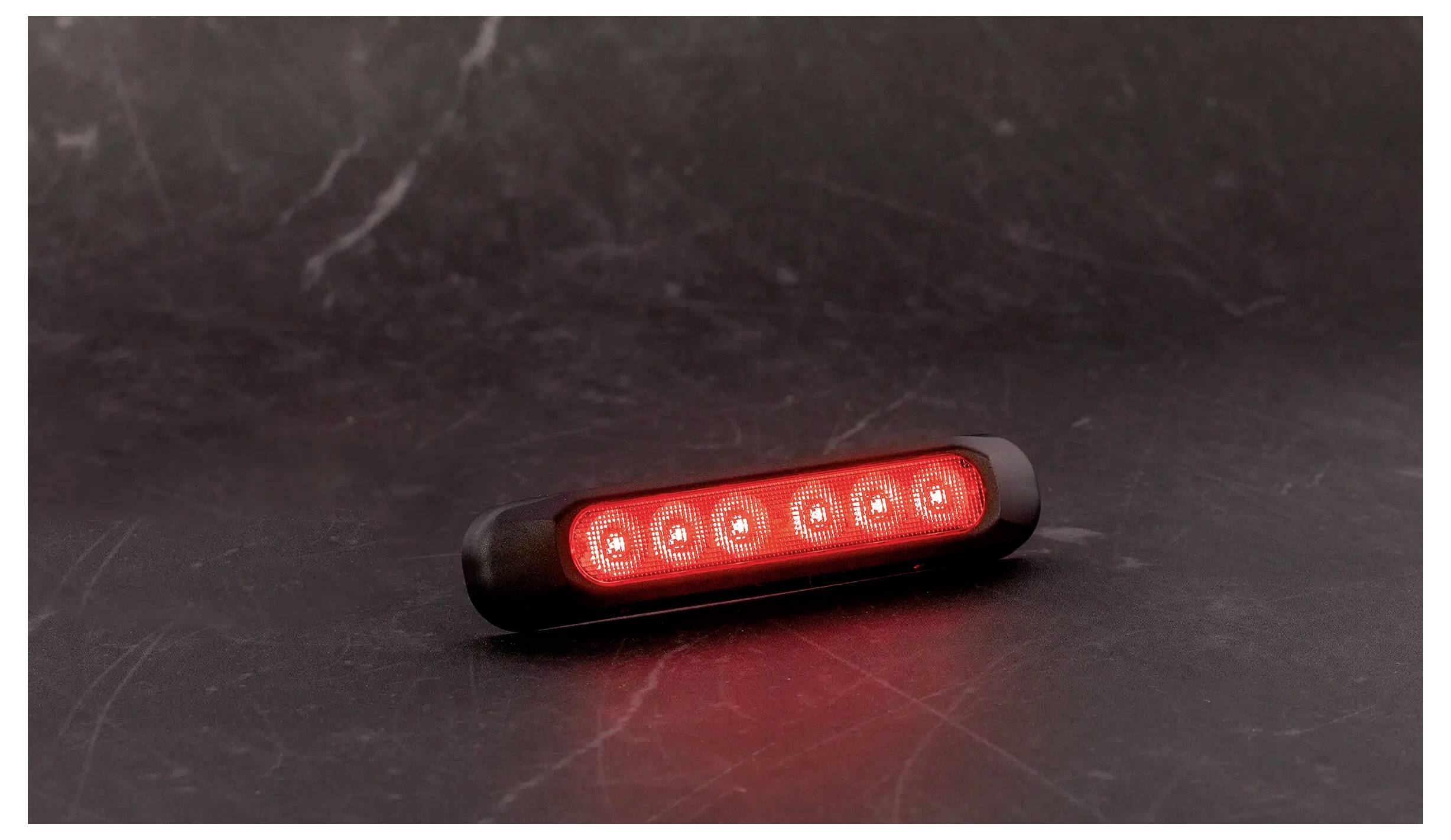 Eine kleine schwarze LED-Leuchte leuchtet rot auf einer dunklen reflektierenden Oberfläche und wirft eine rote Reflexion darunter.
