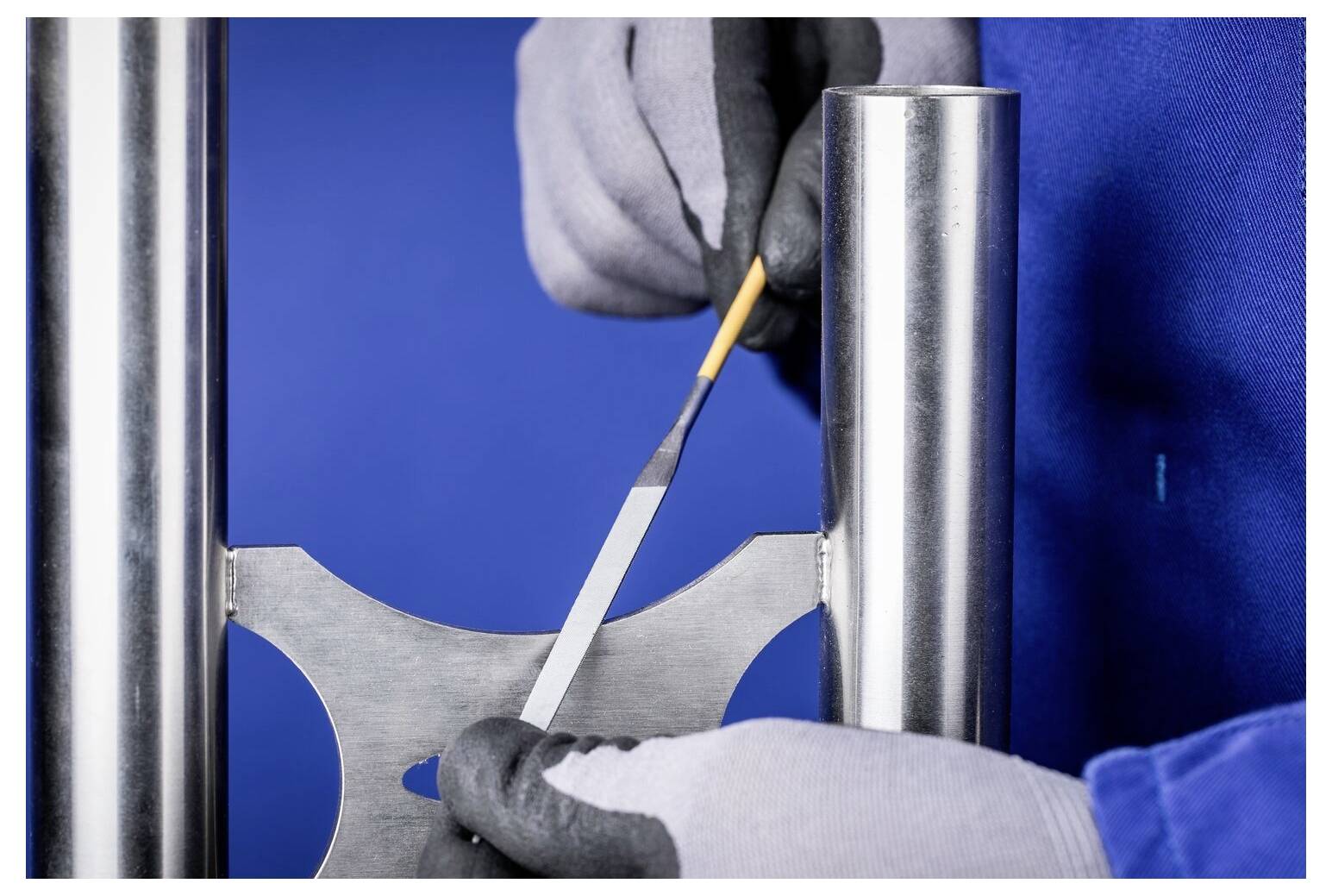 Techniker mit Handschuhen, der ein Metalltestmuster bearbeitet, das zwischen zwei Stahlpfosten gegen einen blauen Hintergrund gehalten wird.