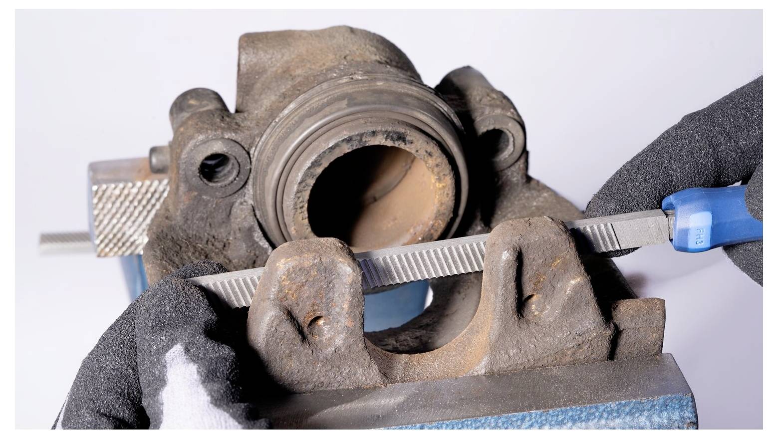 Handschuhe tragende Hände verwenden eine Feile, um Rost von der Innenseite einer in einem Schraubstock eingespannten Bremssattel oder einem ähnlichen Metallteil zu entfernen.