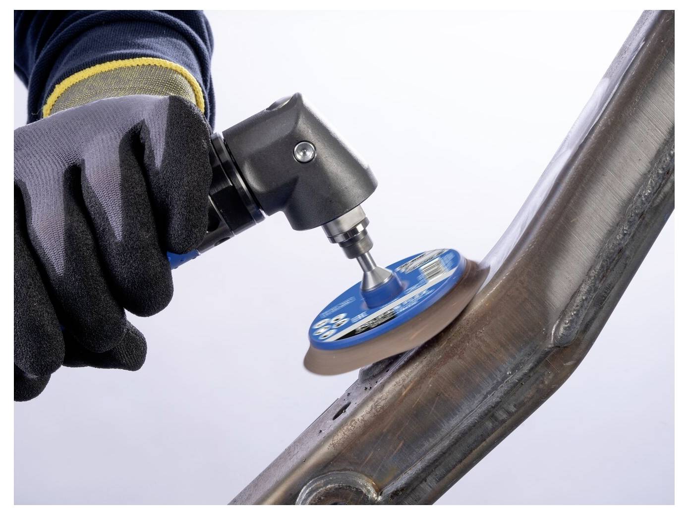 Ein behandschuhter Arbeiter verwendet einen Winkelschleiffer mit einer blauen Schleifscheibe, um eine geschweißte Metallrohrverbindung zu glätten.