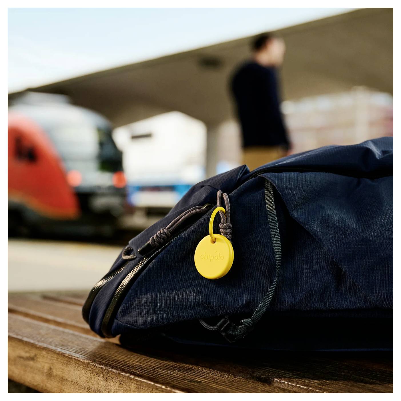 Schwarze Reisetasche mit gelbem Etikett auf einer Bank auf einem Bahnsteig am Flughafen, mit einem Zug und einer unscharfen Person im Hintergrund.