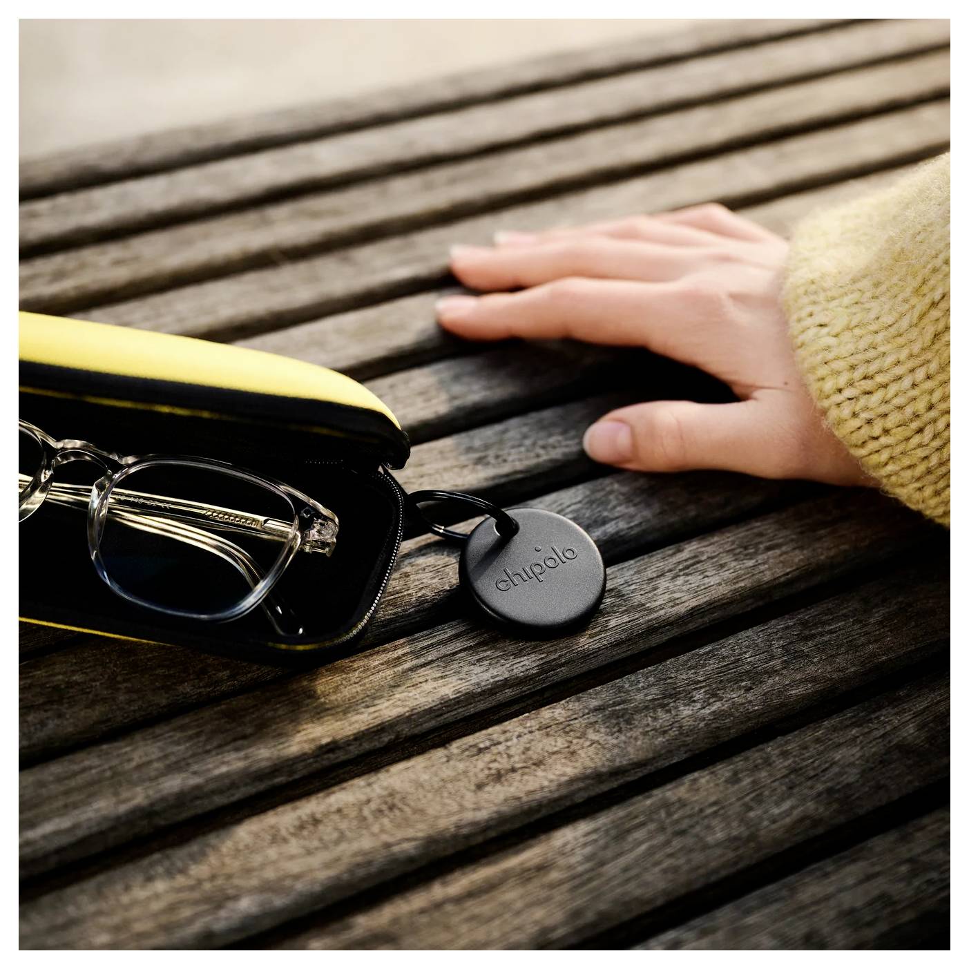 Eine Hand greift nach einer Brille in einem gelben Etui mit einem Chipolo-Bluetooth-Tracker auf einer Holzbank.
