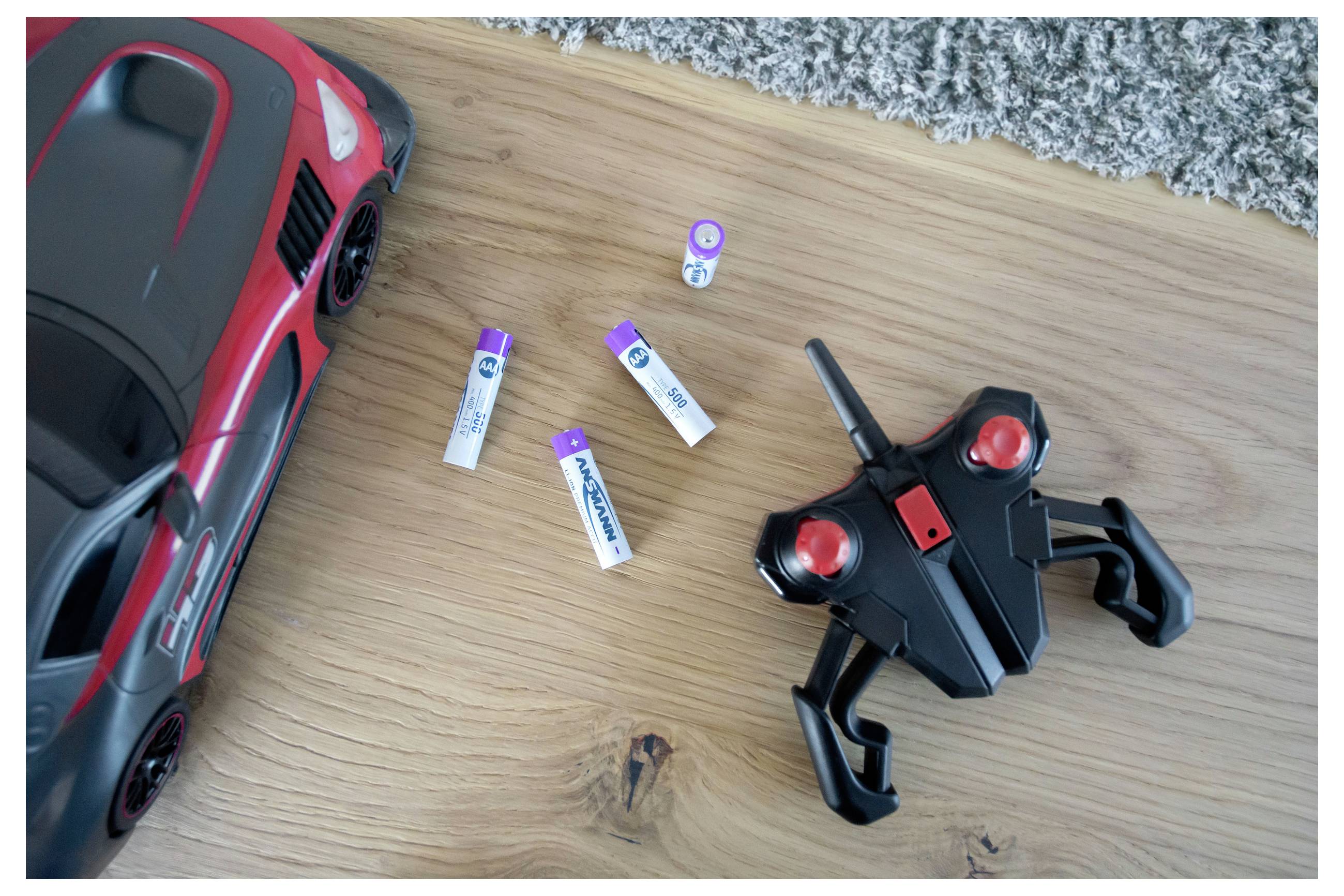 Rotes Spielzeugauto und schwarzer Fernbedienung neben mehreren AAA-Batterien auf einem Holzboden