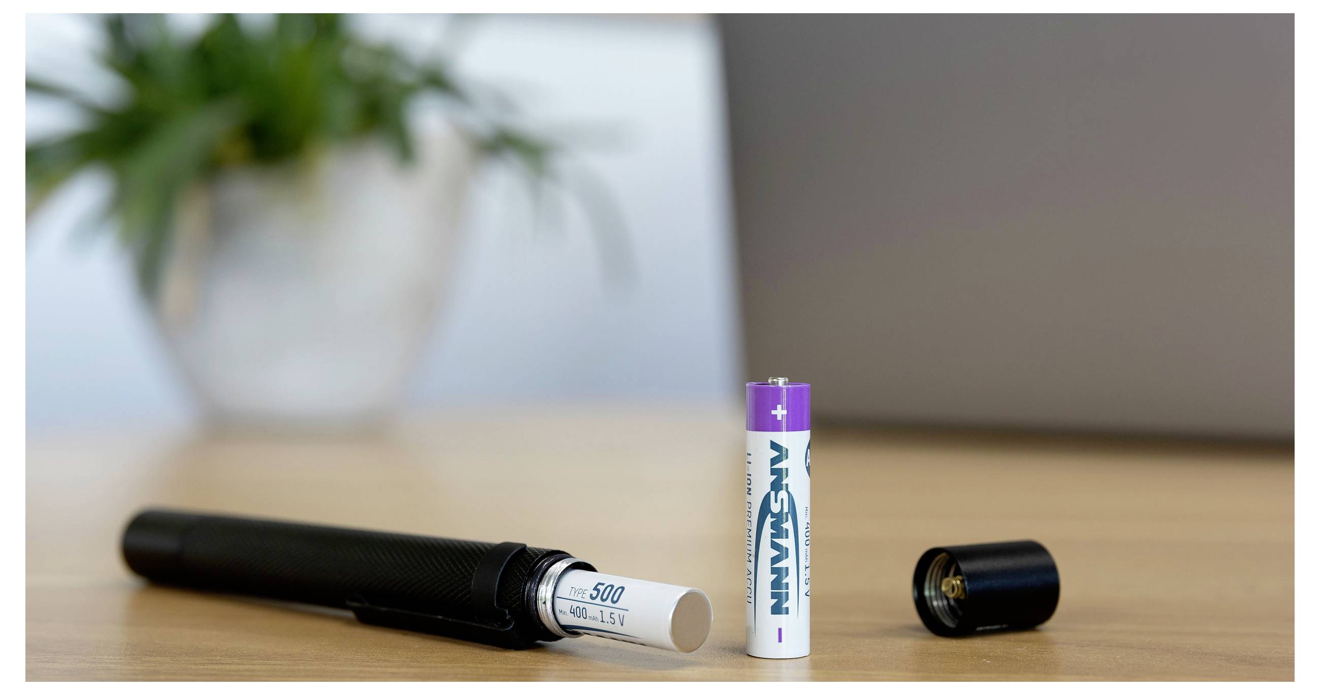 Zwei AA-Batterien und ein schwarzes zylindrisches Gerät liegen auf einem Holztisch neben einem Laptop, was auf einen batteriebetriebenen Stift oder ein Gadget mit entfernter Kappe hindeutet.