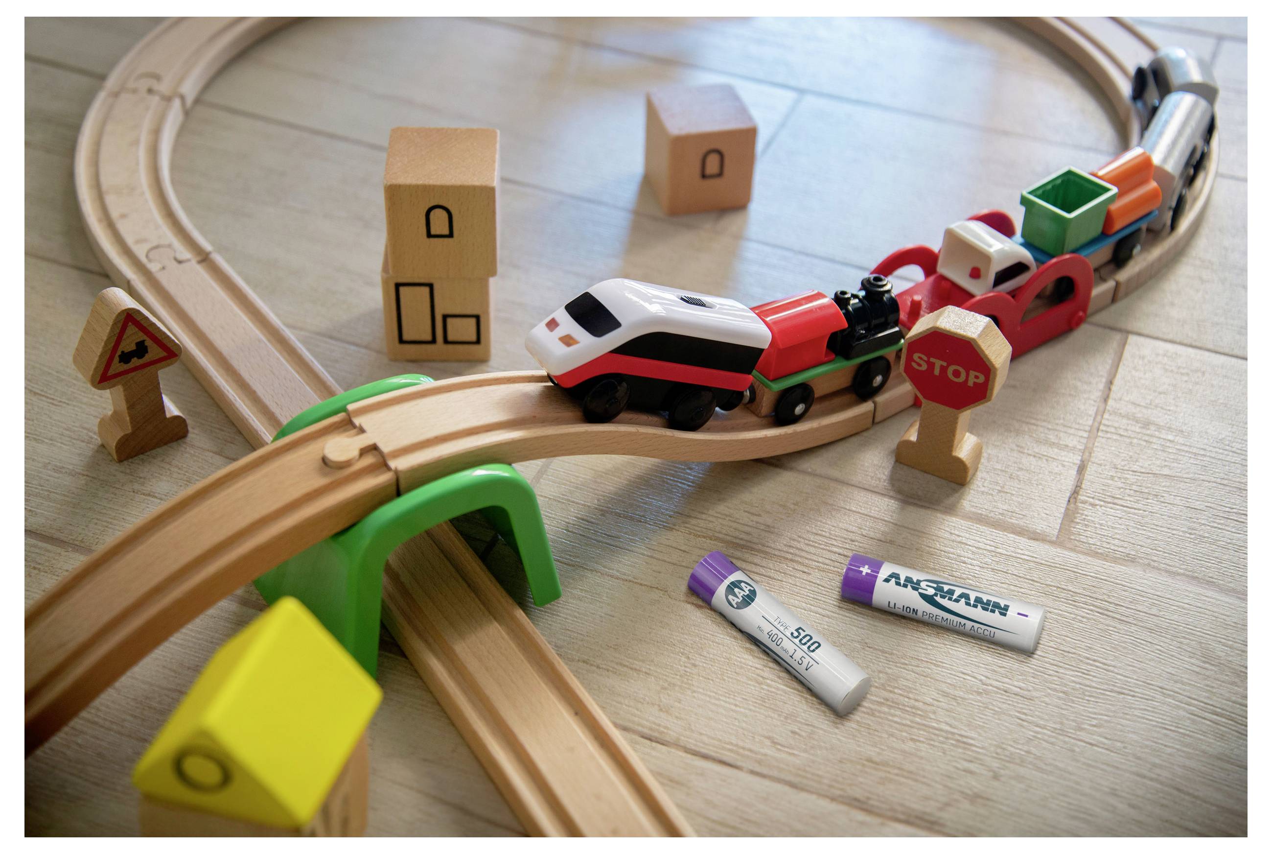 Spielzeugeisenbahn und Autos auf einer Holzbahn mit Stoppzeichen, Pappgebäuden und verstreuten Batterien in der Nähe auf dem Boden.