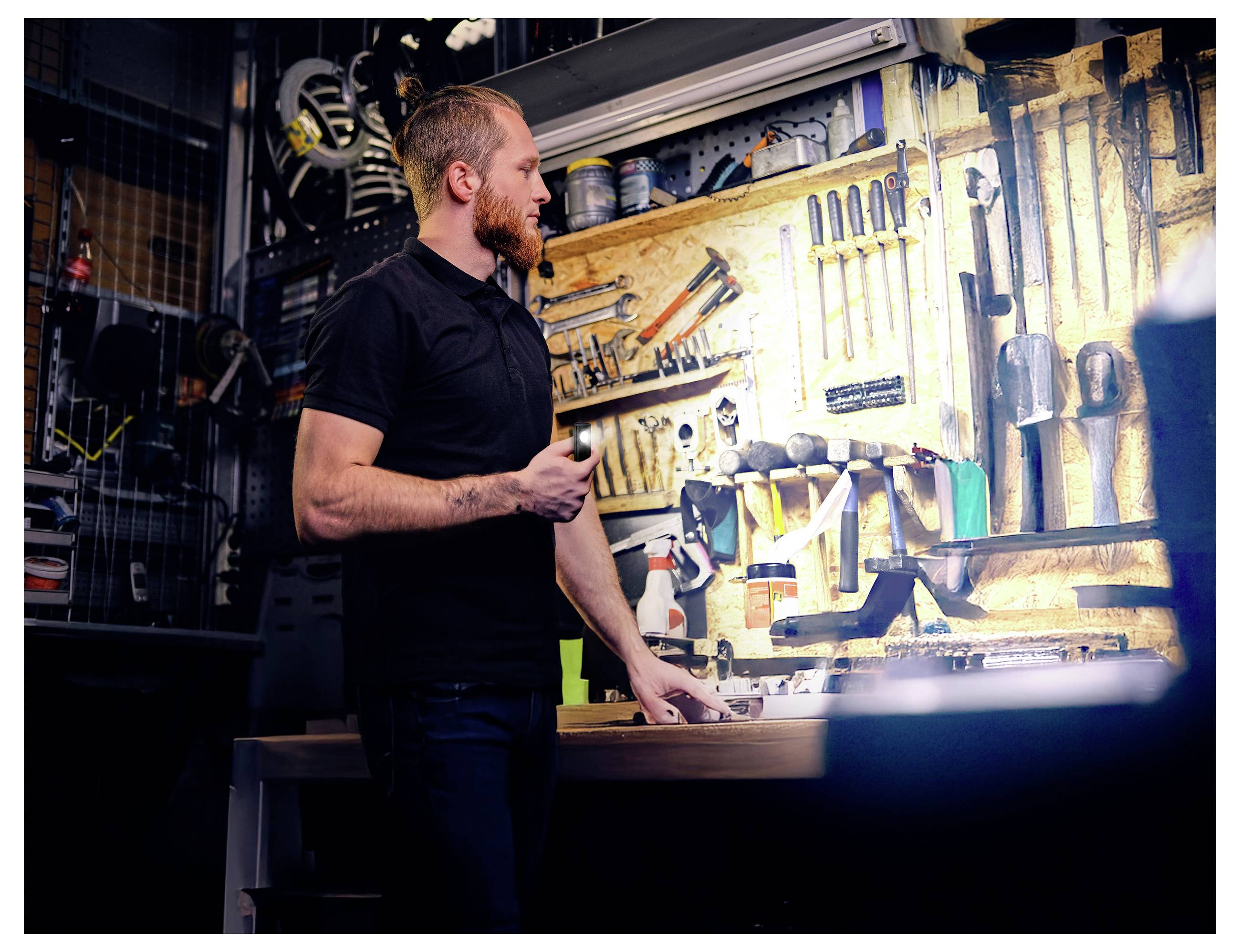 Ein Mann in einer Werkstatt steht an einer Werkbank, hält eine Taschenlampe und untersucht Handwerkzeuge, die an einer Wand montiert sind.