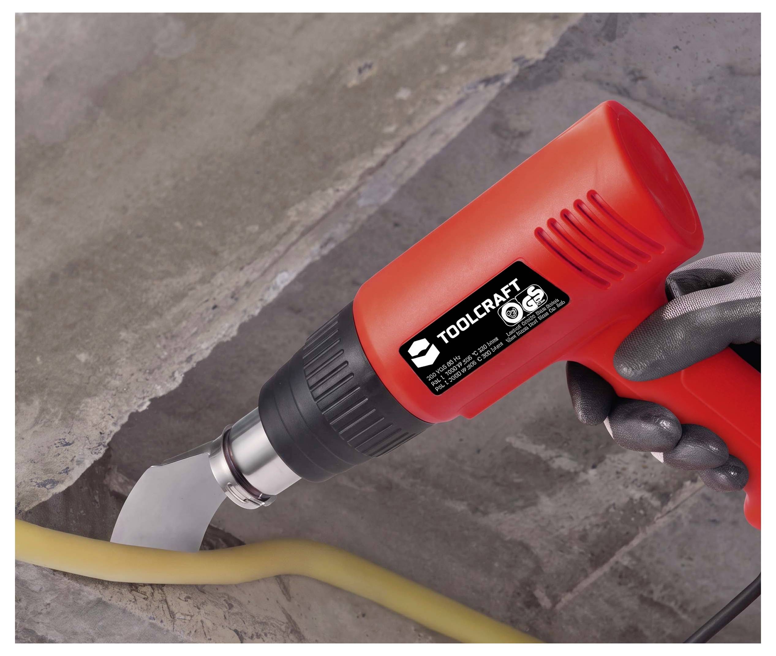 Eine behandschuhte Hand benutzt eine rote Toolcraft-Heißluftpistole, um ein gelbes Kabel auf einem Betonfußboden zu biegen oder zu erhitzen.