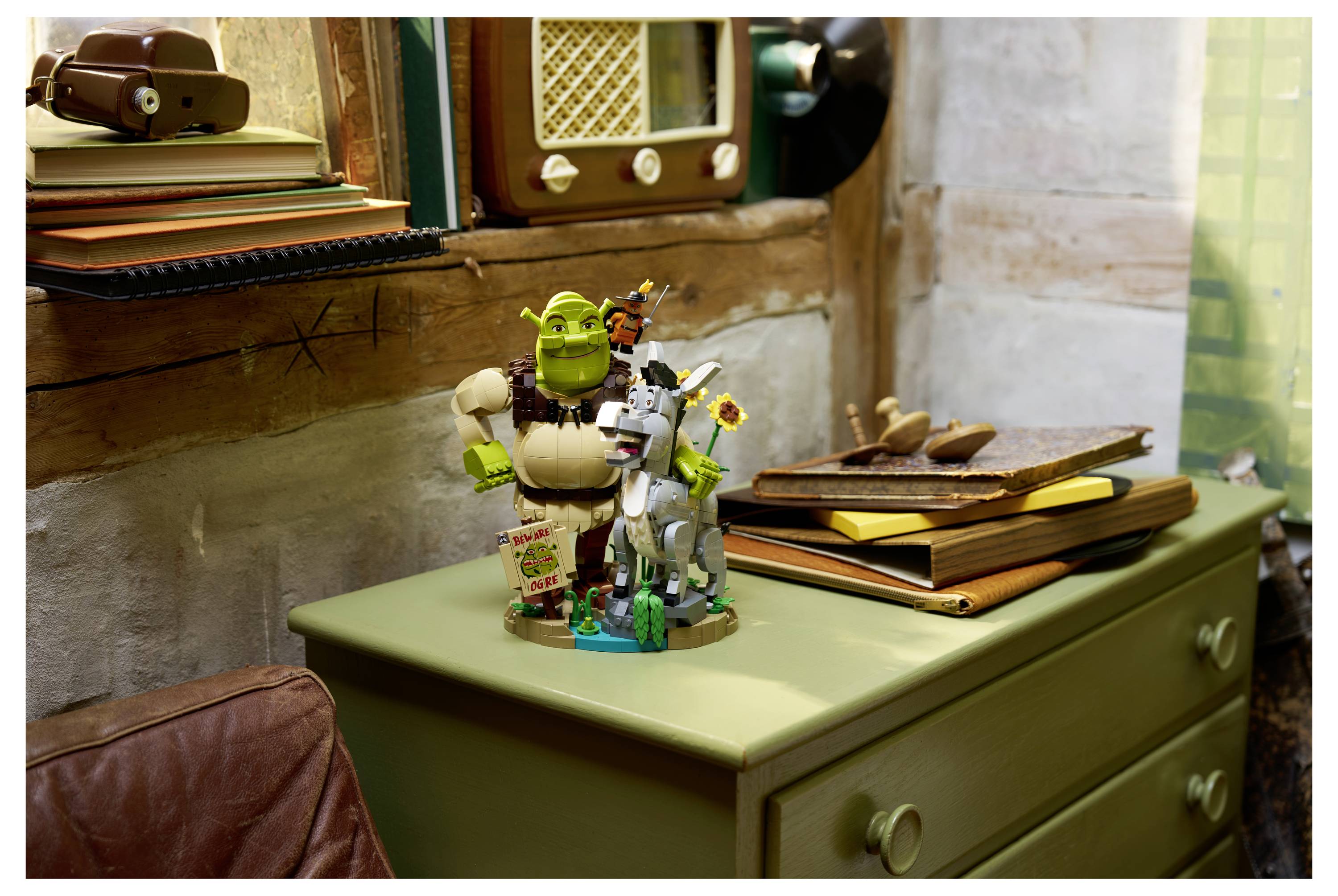 Spielzeugfiguren von Shrek und einem Esel stehen auf einer grünen Kommode neben gestapelten Büchern in einem gemütlichen Zimmer.