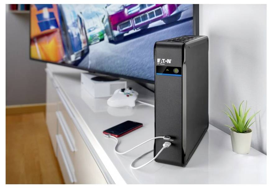 Eaton-Backup-Stromversorgungsgerät auf einem weißen Fernsehständer, an ein Smartphone angeschlossen und neben einem Spielcontroller, mit einem Fernseher und einer kleinen Pflanze im Hintergrund.