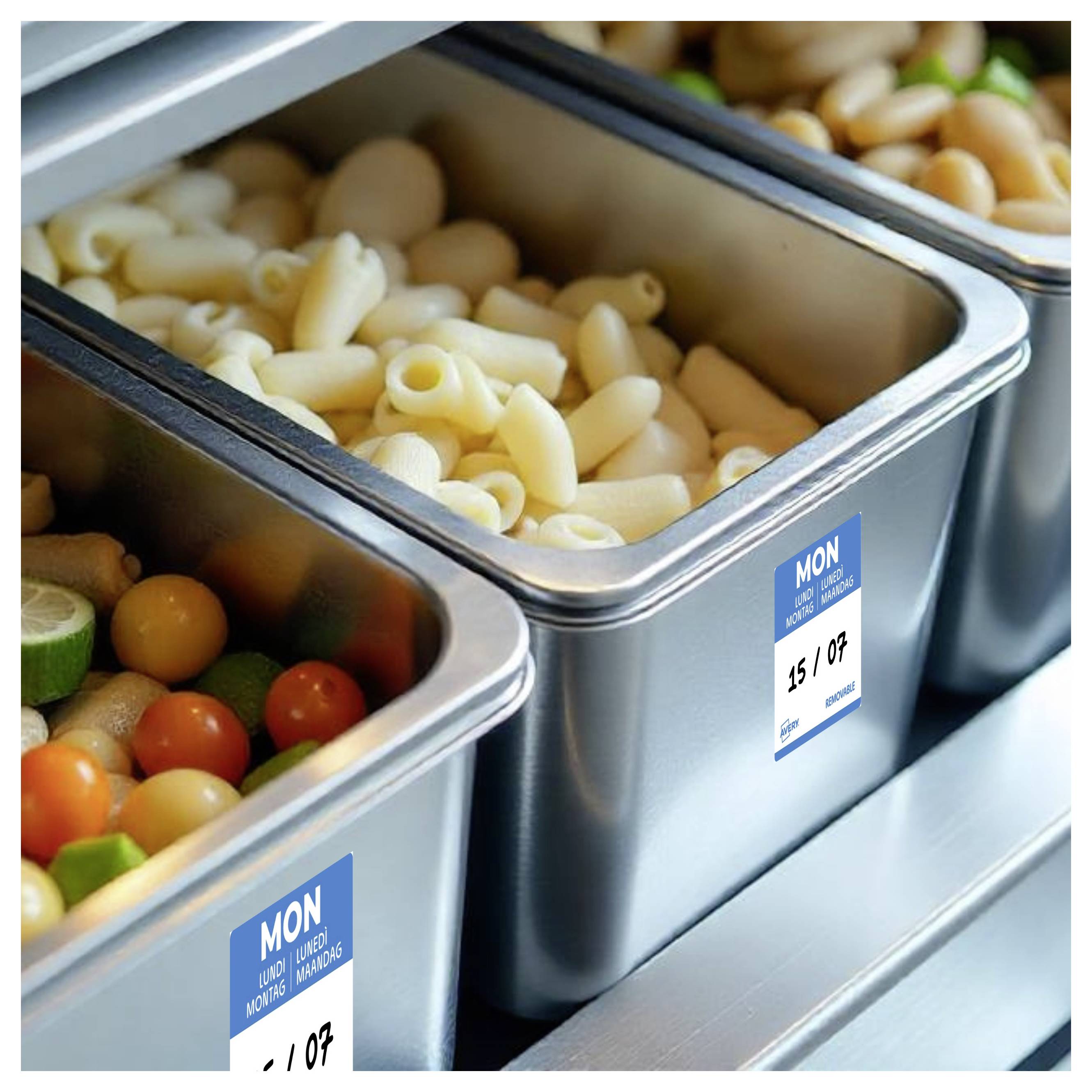 Eine Cafeteria-ähnliche Lebensmittelzubereitungsstation mit Edelstahlpfannen, die gekochte Nudeln und frisches Gemüse enthalten, gekennzeichnet mit blauen Datumsetiketten.