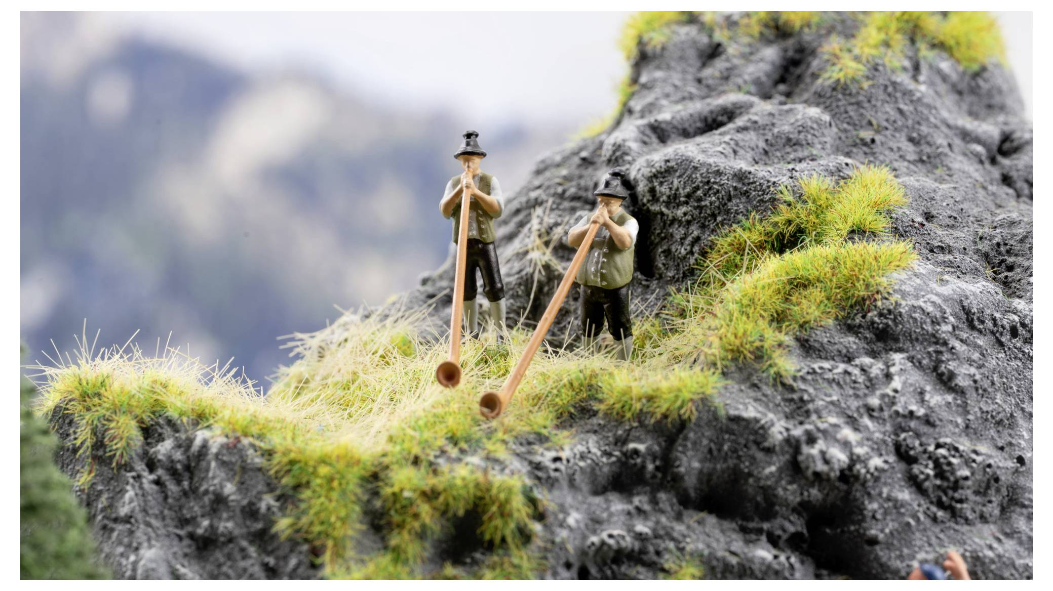 Zwei Wanderer spielen lange Holzdidgeridoos auf einem felsigen Berghang mit Gras vor einem unscharfen Alpenaufnahmen Hintergrund.