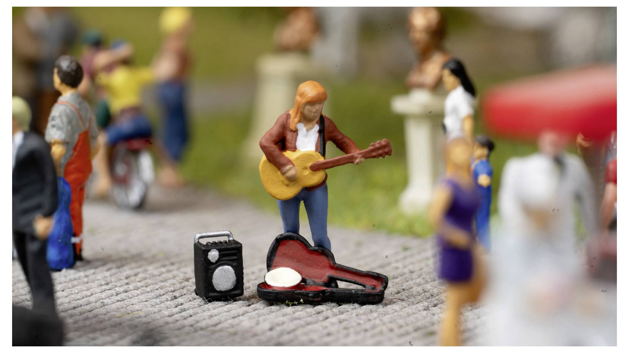 Kleine Straßenmusiker-Figur, die neben einem offenen Instrumentenkoffer und einem kleinen Verstärker Gitarre spielt, mit unscharfen Fußgängern im Vorder- und Hintergrund.