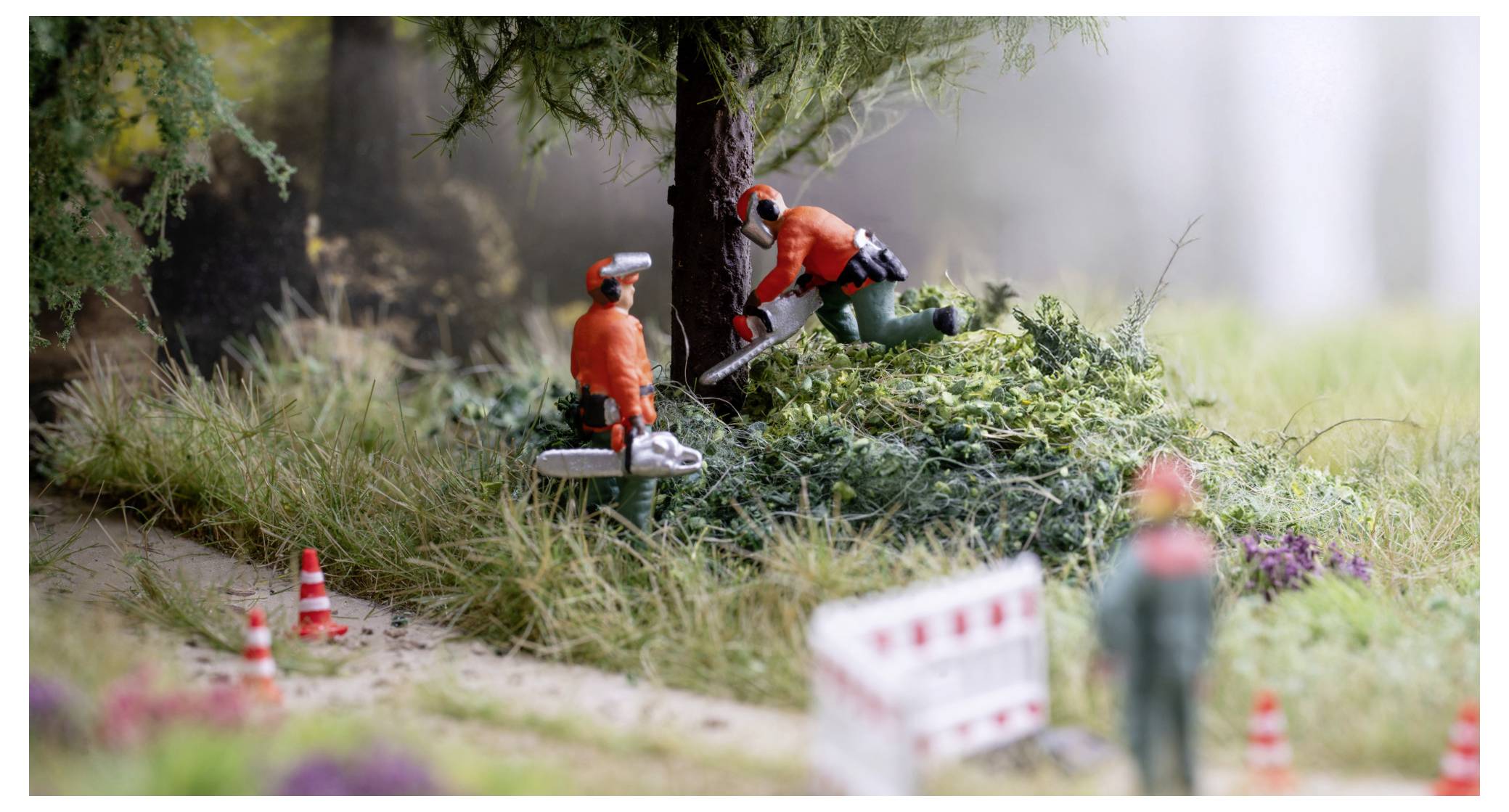 Miniatur-Straßenbaustelle mit zwei Arbeitern in Orange, die Gestrüpp um einen Baum herum entfernen, mit Verkehrskegeln und Absperrungen entlang eines Weges.