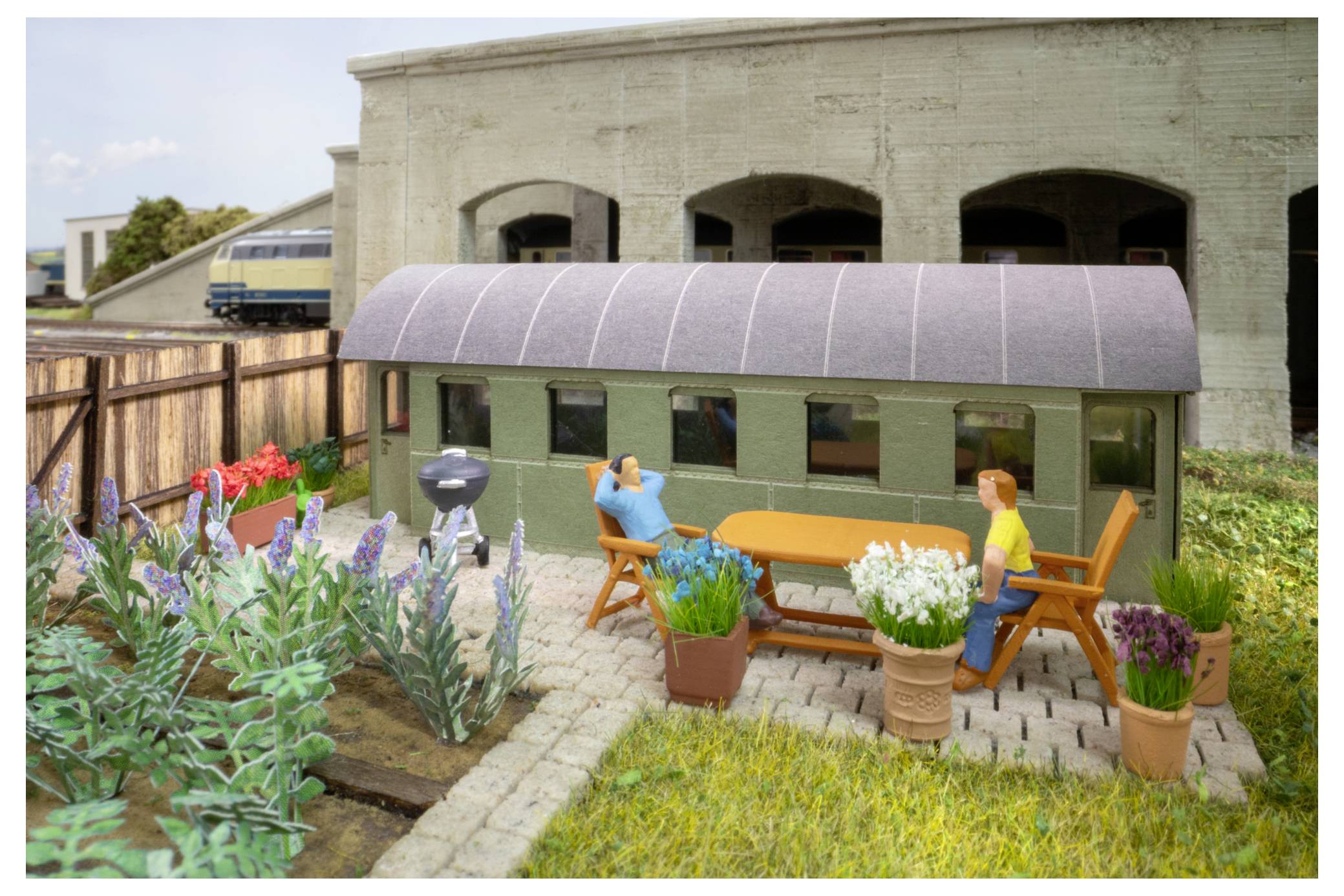 Miniatur-Gartenterrassen-Szenerie mit zwei sitzenden Figuren an einem orangefarbenen Tisch neben Topfblumen, einem Grill und einem gewächshausähnlichen Schuppen.