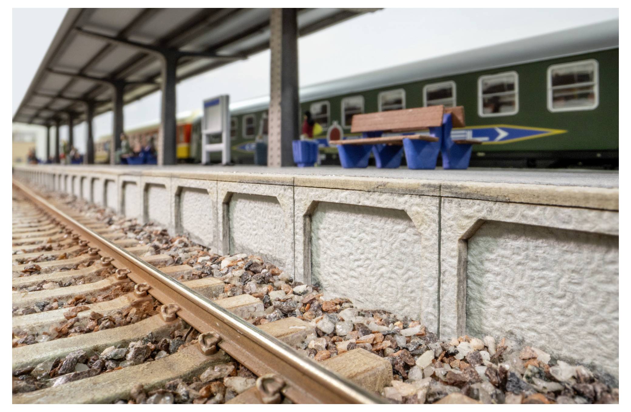 Modelleisenbahnplattform neben einem Eisenbahngleis, mit einem grünen Personenwagen und einer Bank unter einem überdachten Bahnhofsdach.