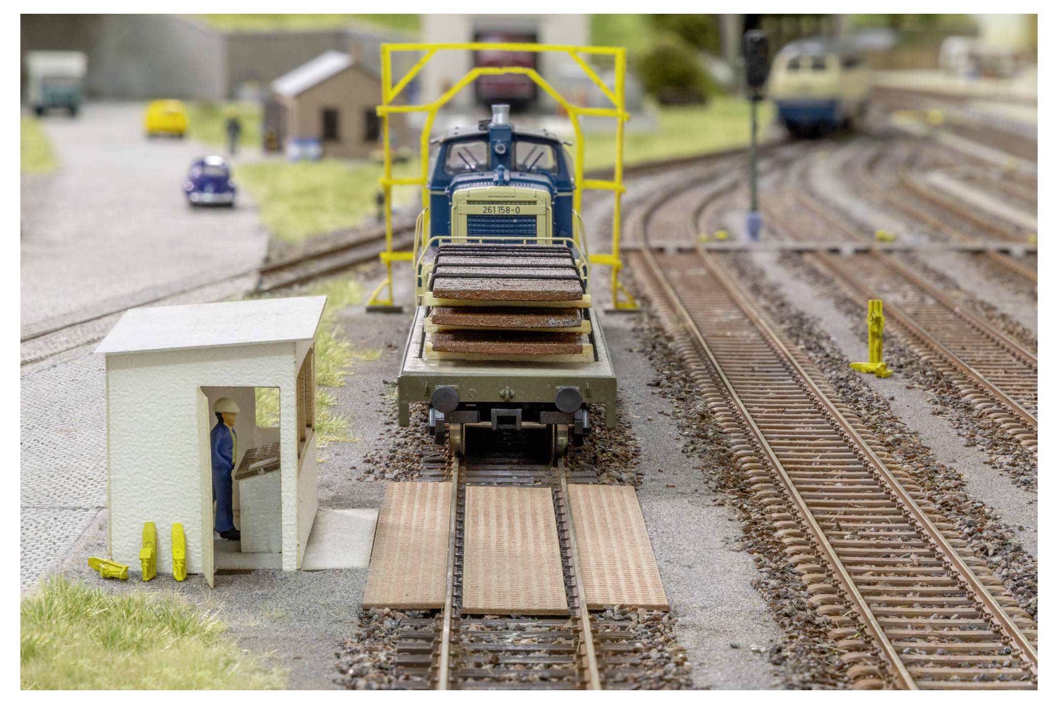 Modelleisenbahn auf Gleisen, die sich einer kleinen Eisenbahnkreuzung nähert, mit einem Miniaturarbeiter neben einem Gleis-Schuppen und mehreren Eisenbahnlinien im Hintergrund.