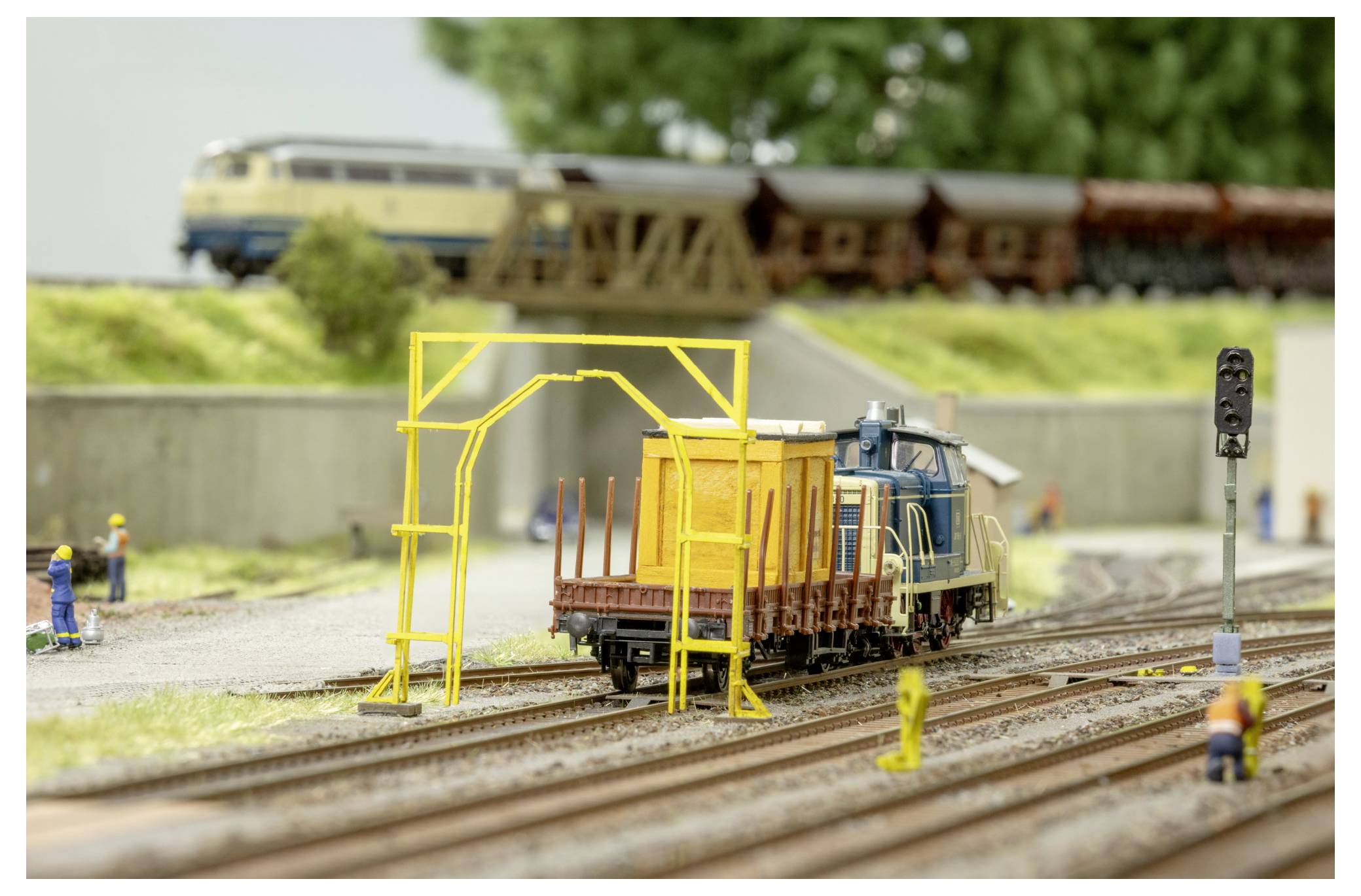 Modelleisenbahn-Szenerie mit einer blauen Lokomotive, die Güterwagen durch einen gleiseitigen Wartungshof zieht, mit Miniaturarbeitern und einem gelben Portalkran.