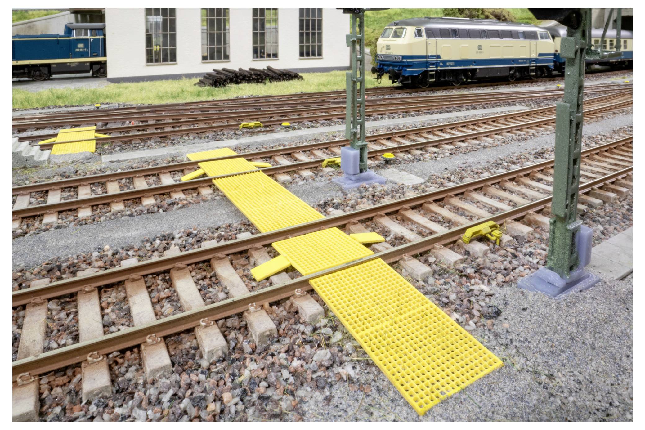 Modelleisenbahnhof mit mehreren Gleisen und leuchtend gelben Fußgängerübergängen über den Schienen, mit einer blau-creme farbenen Lokomotive im Hintergrund.