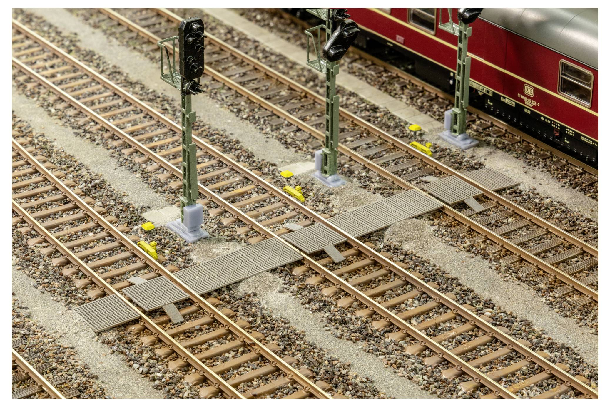 Modelleisenbahnübergang mit mehreren Gleisen, Signalmasten und einem roten Personenzug, der oben rechts teilweise sichtbar ist.